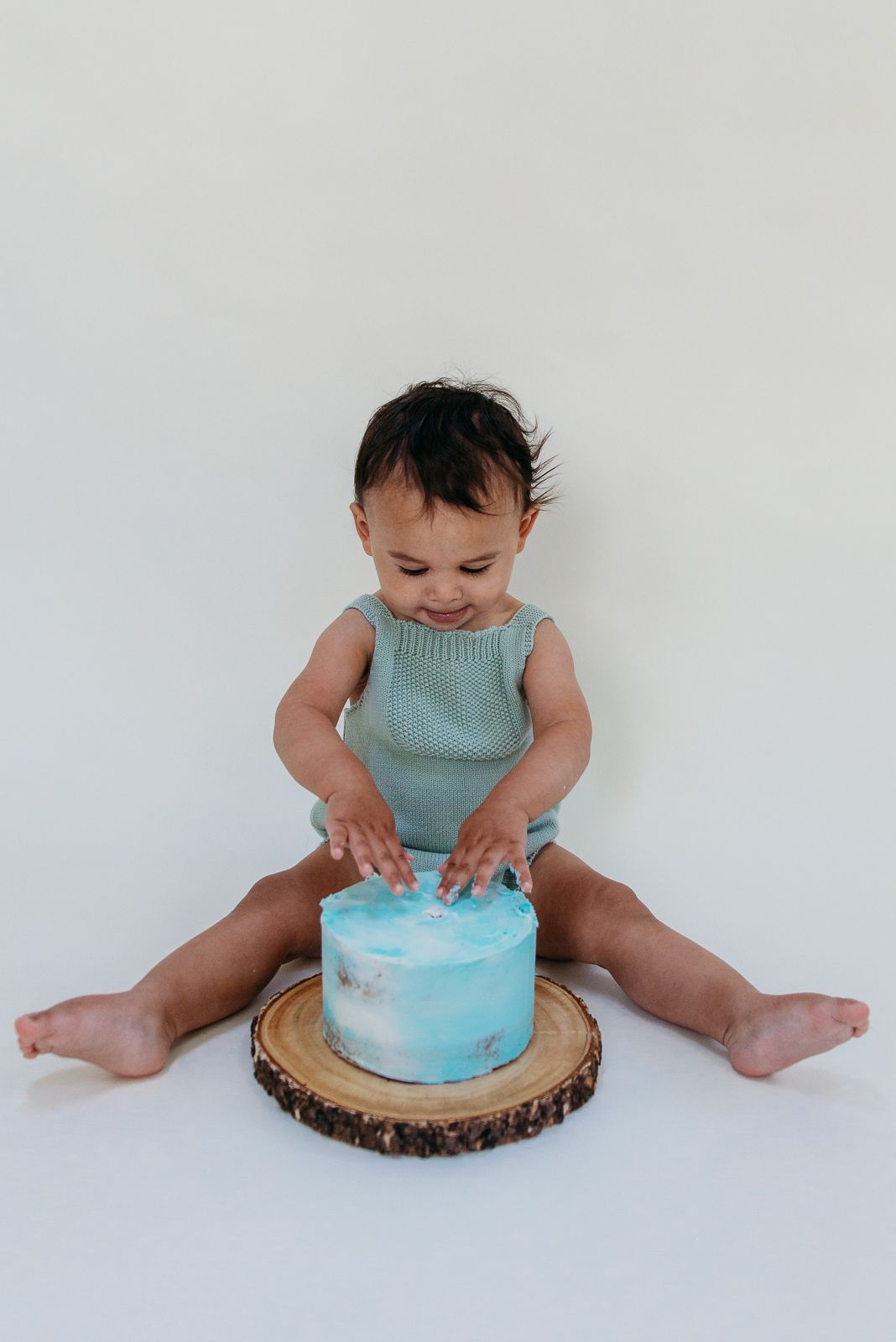 Baby in mint romper playing with blue and white cake on wooden platter.