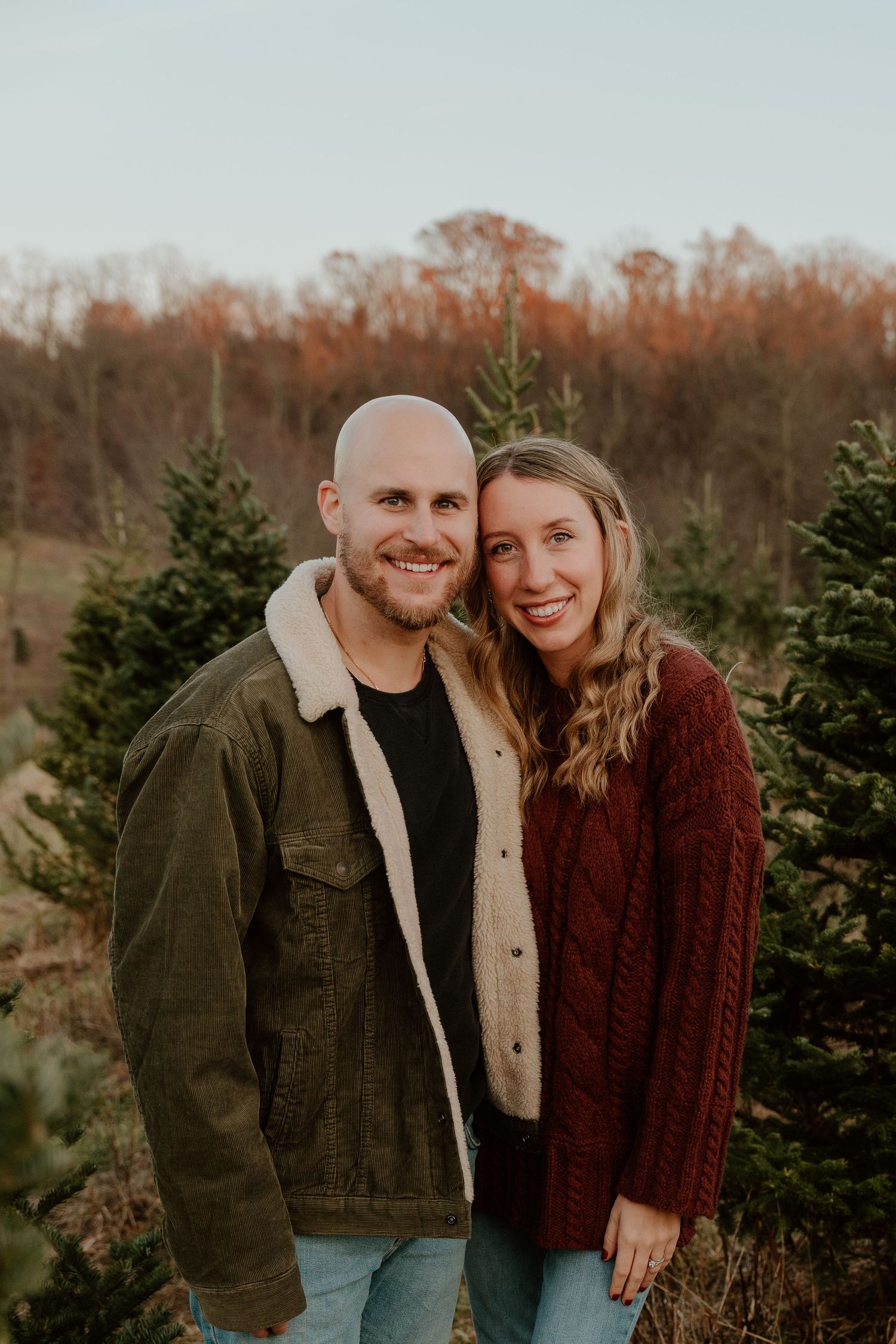 A couple poses together at a Christmas tree farm during sunset wearing cozy winter attire.