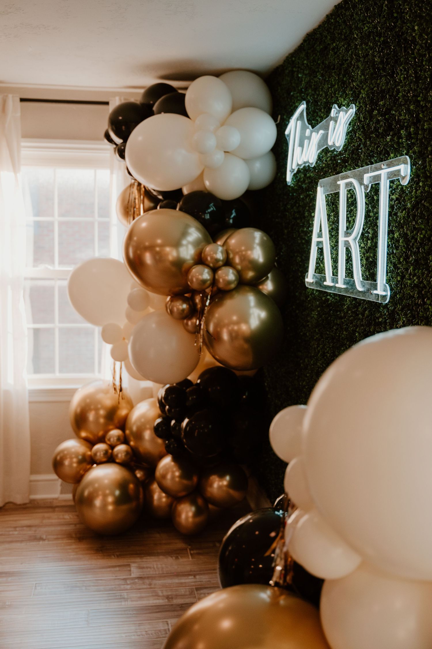 Modern party decor featuring an elegant balloon arch with black, gold and white balloons against a dark green backdrop.