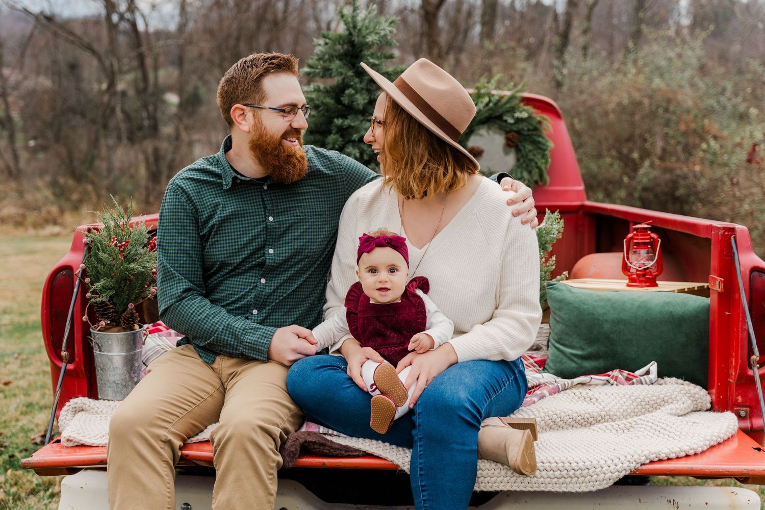Family sitting in red truck bed with evergreen trees and holiday decor.