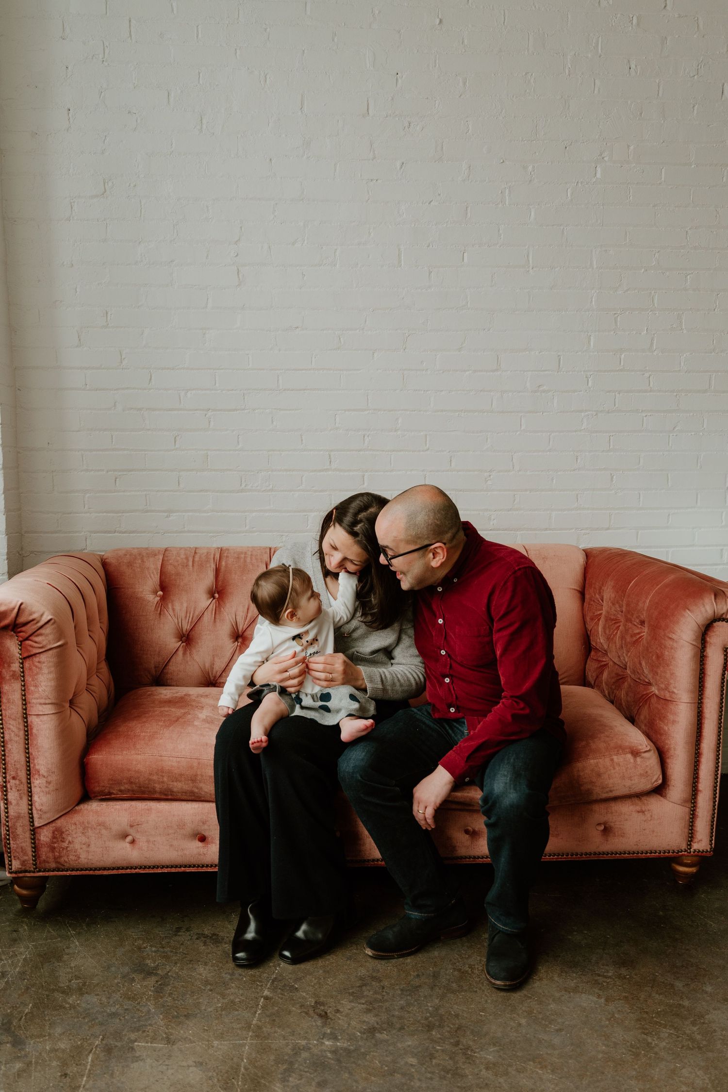 A family sits together looking at something on a pink vintage tufted sofa against a white brick wall.