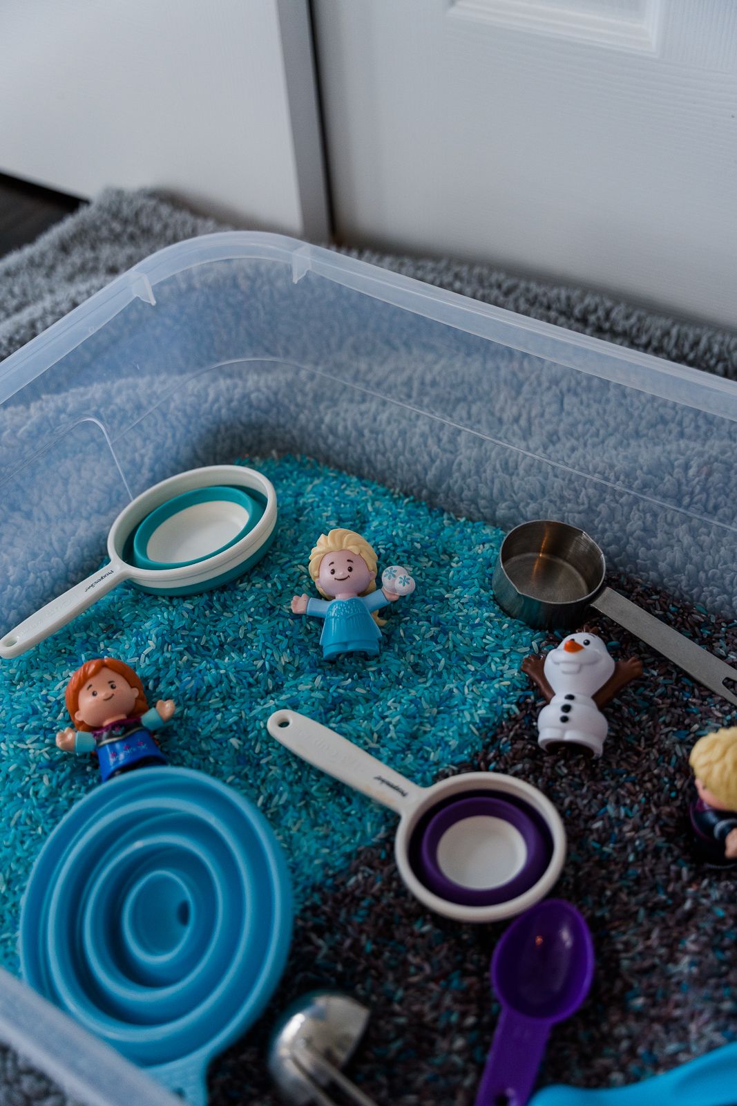 Frozen-themed sensory bin with blue rice and character figurines alongside measuring cups.