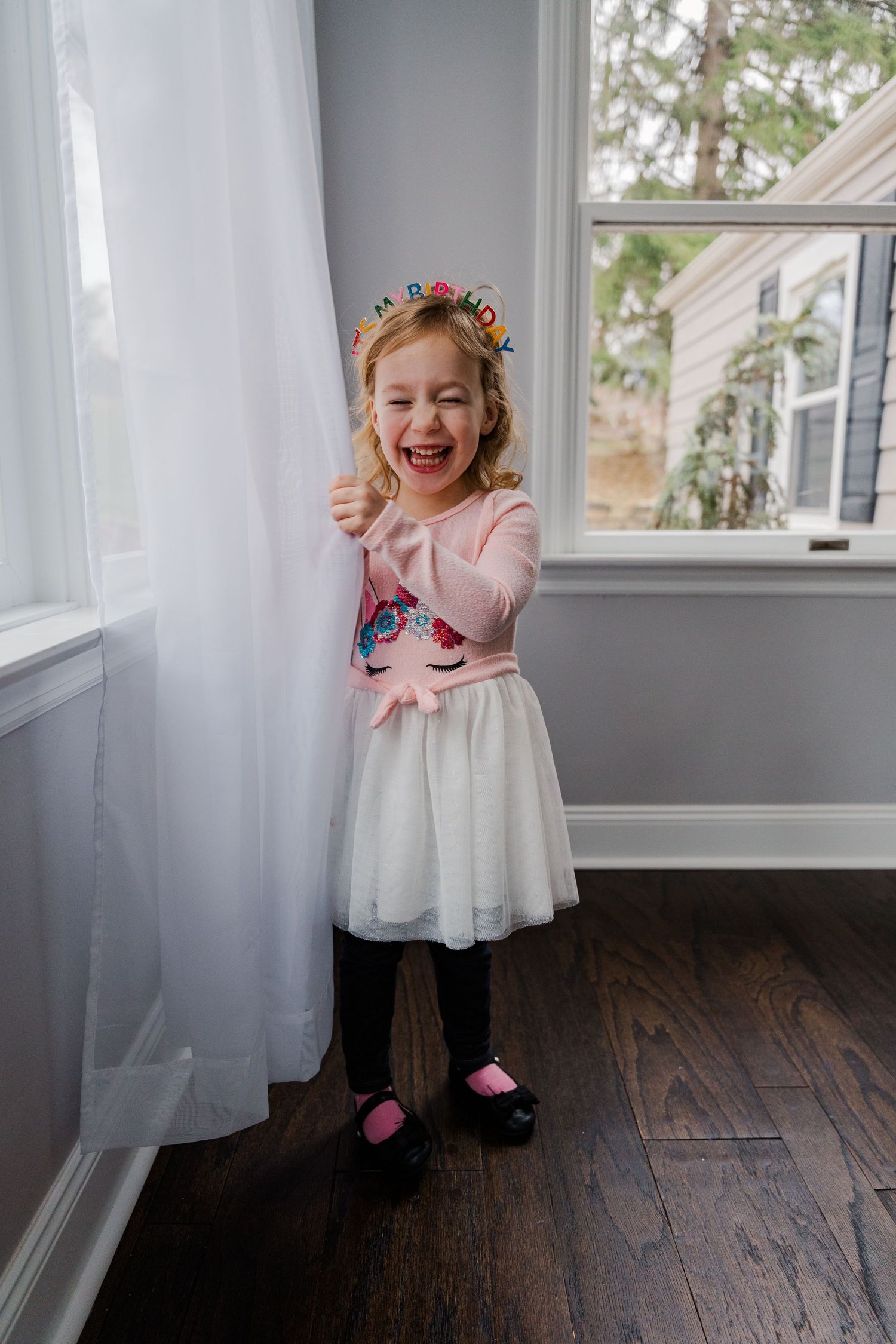 Young child in tulle dress and flower crown laughing by window.