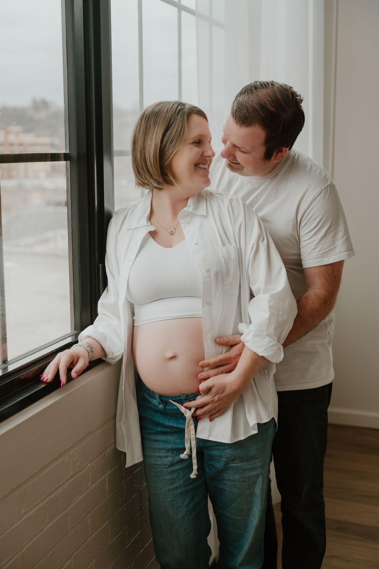 Intimate maternity photo by a window shows expecting couple in white tops sharing tender moment together.