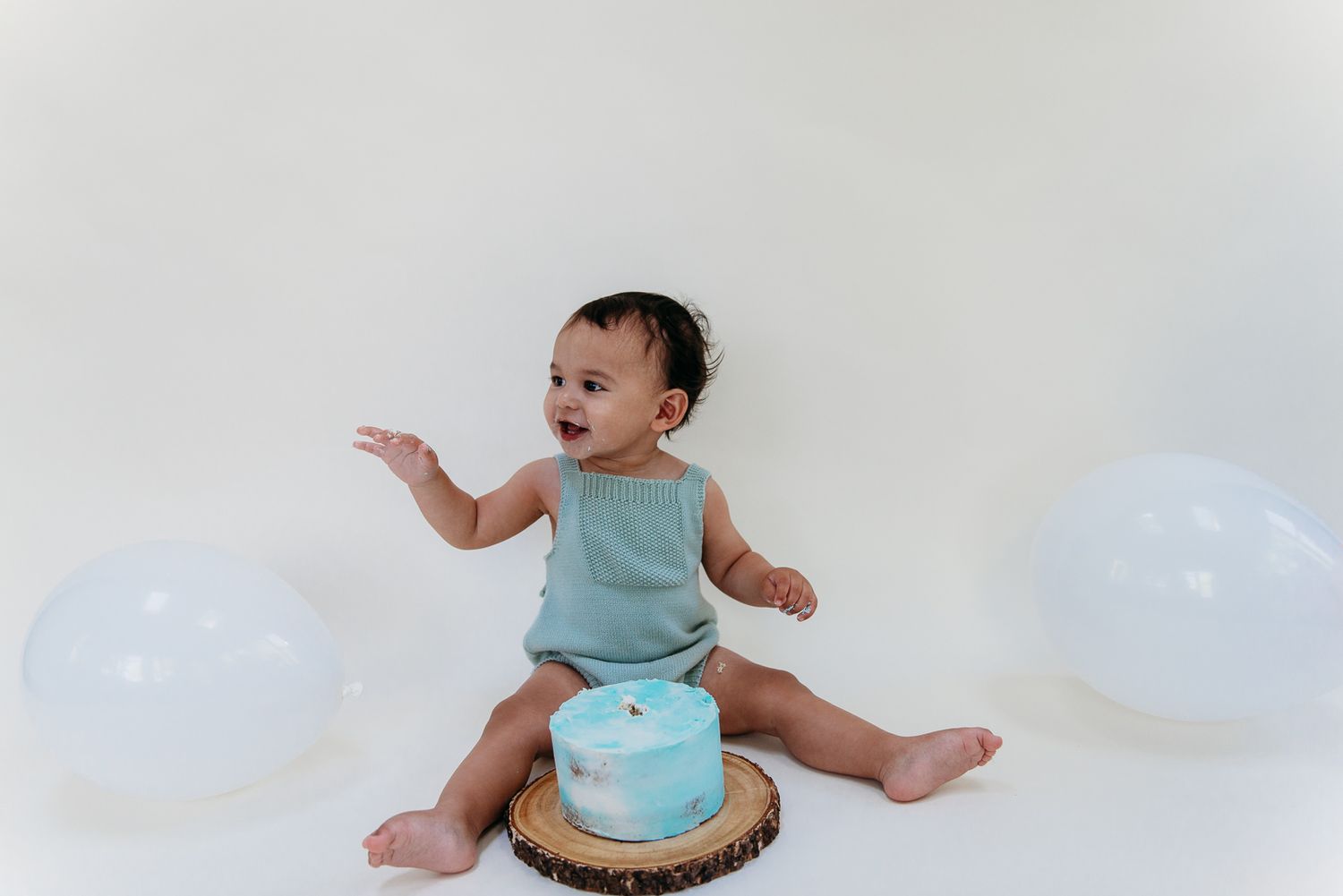 Baby in mint outfit celebrating with cake and white balloons.