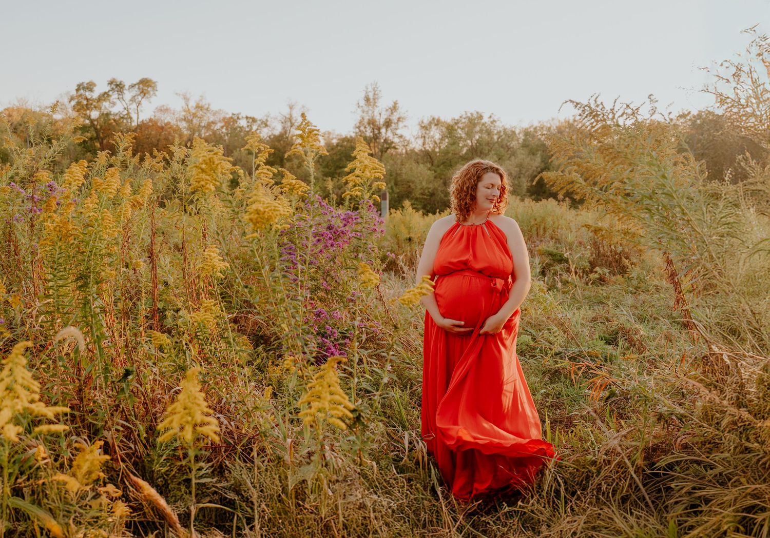 A figure in a flowing red dress stands gracefully among wildflowers and tall grass during golden hour.
