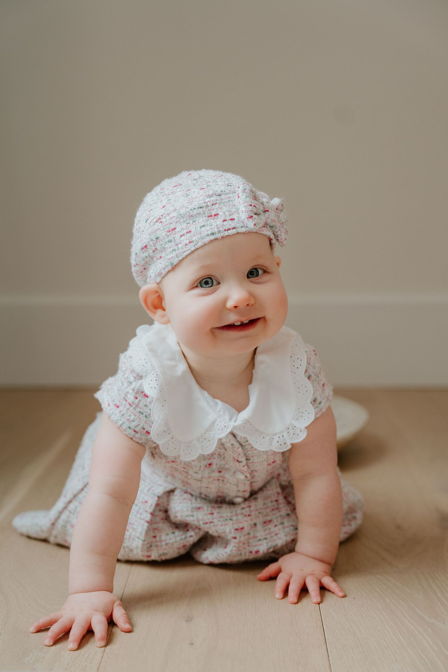 A baby wearing a floral dress and matching bonnet crawls on a light wooden floor with a sweet smile.