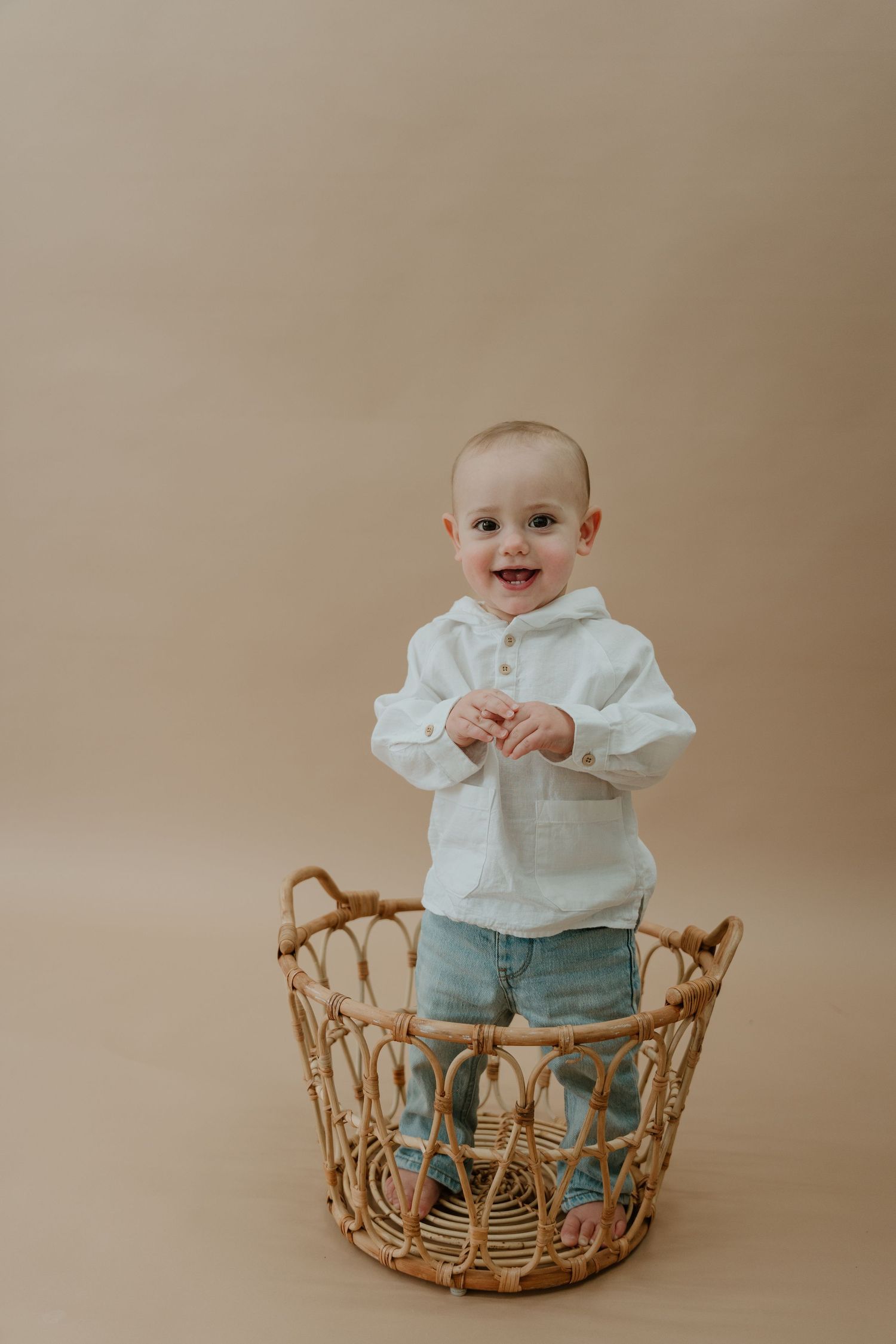 A small child stands in a woven rattan basket wearing a white shirt and jeans against a neutral beige background.