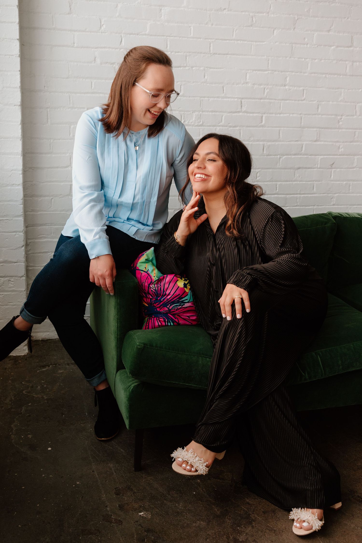 Two friends share a joyful moment while sitting on a dark green couch against a white brick wall.