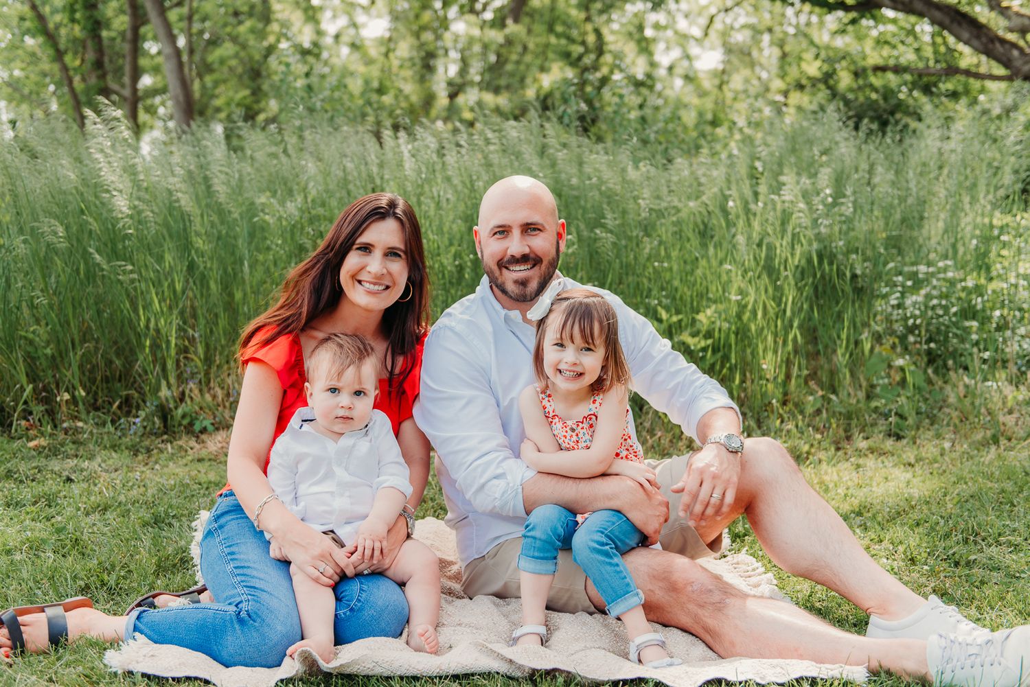 A family sits together on a blanket outdoors surrounded by tall green grass on a sunny day.