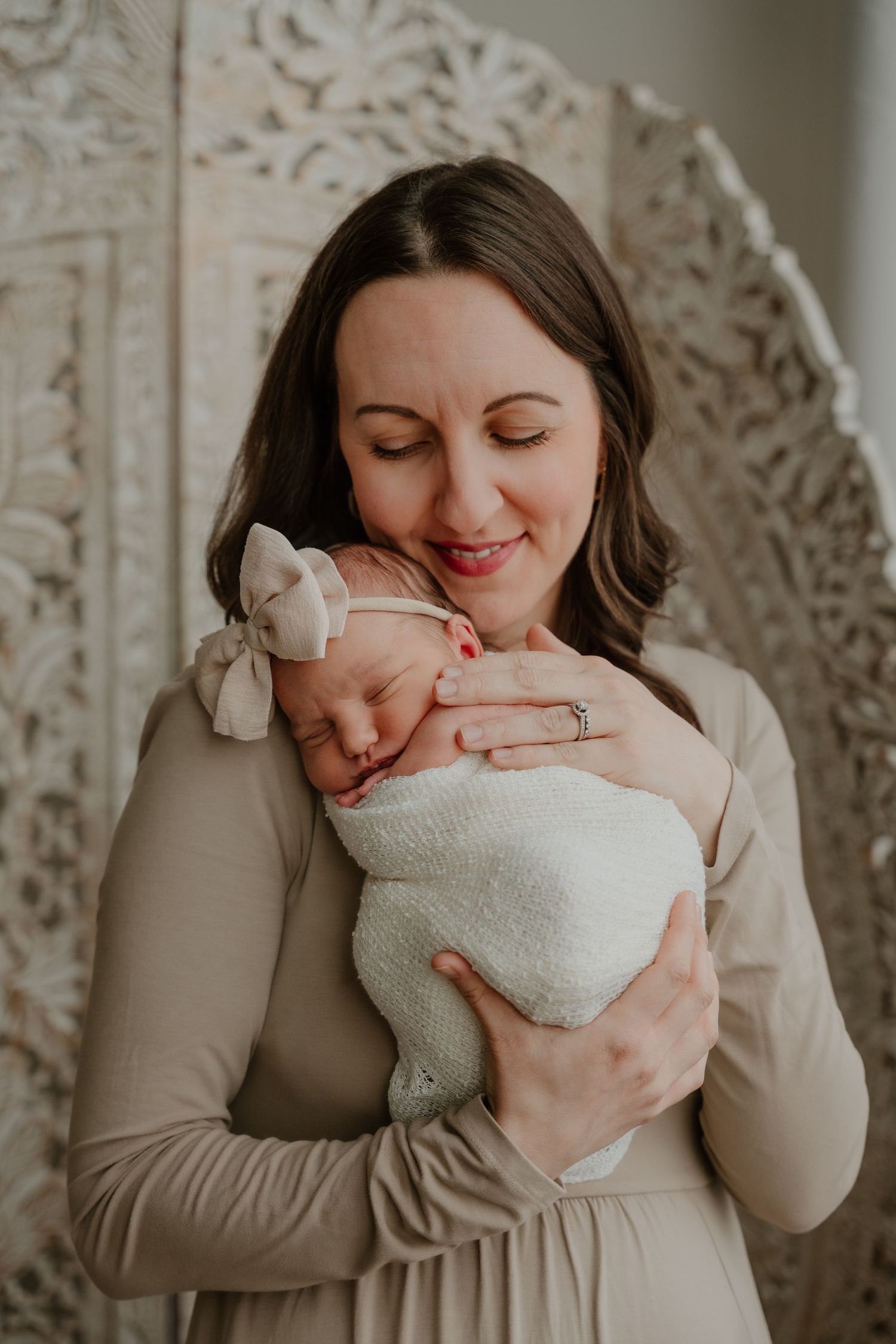 A tender moment captured of a woman in beige dress holding and gazing lovingly at a sleeping newborn swaddled in white.