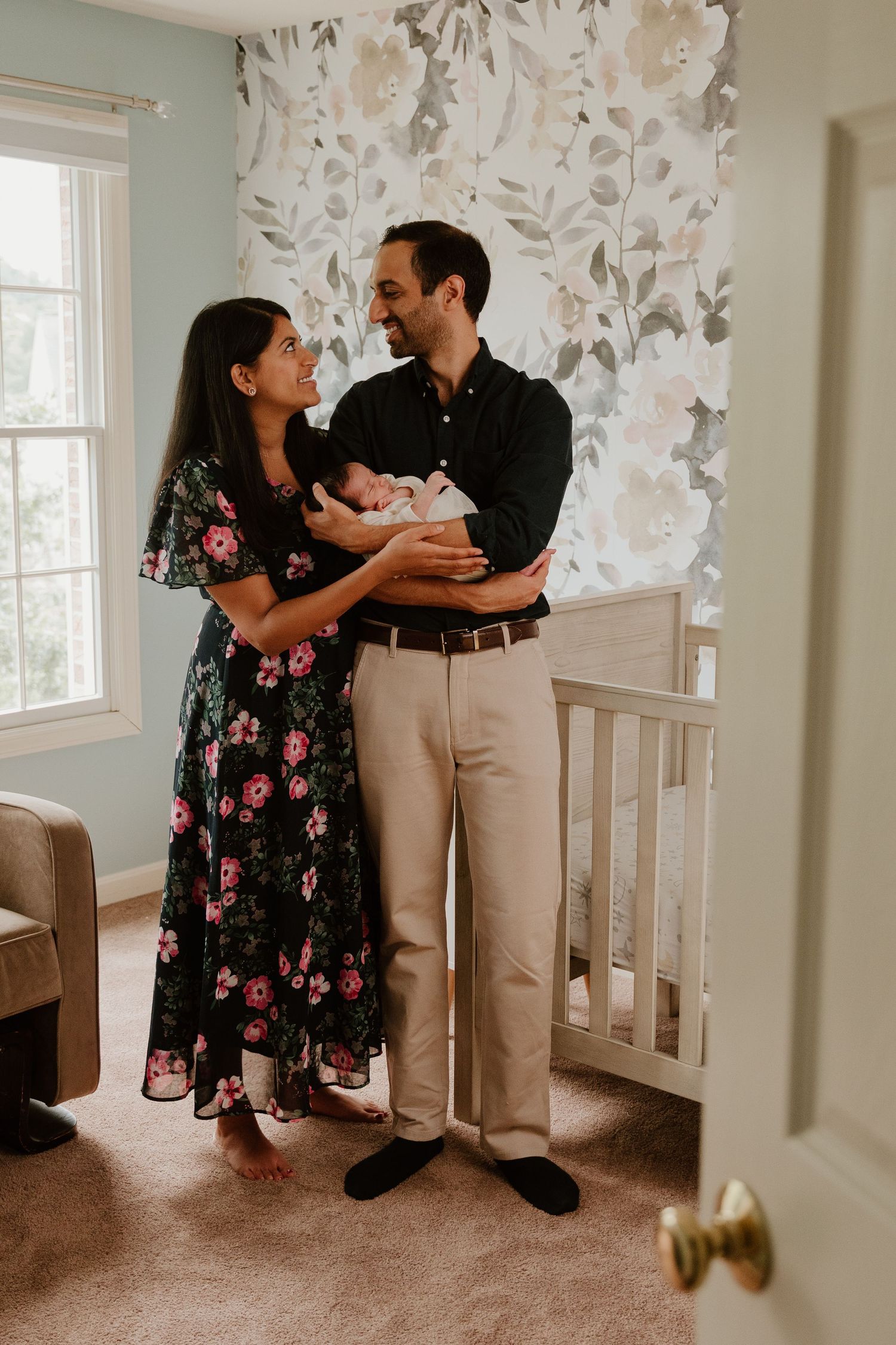 New parents share an intimate moment with their newborn baby in a nursery with floral wallpaper.