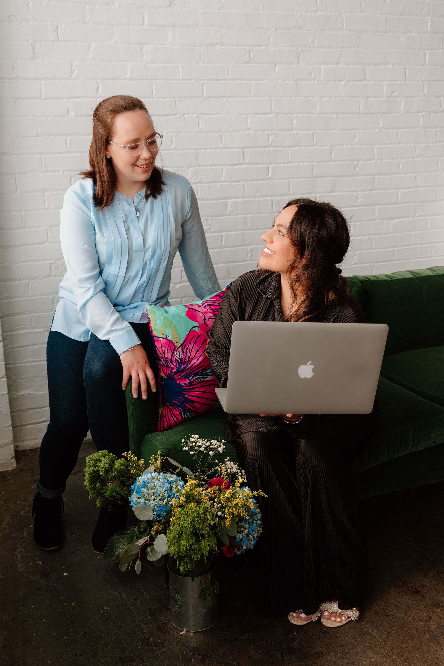 Two people in casual attire interact playfully with a laptop while sitting near a floral arrangement against a white wall.