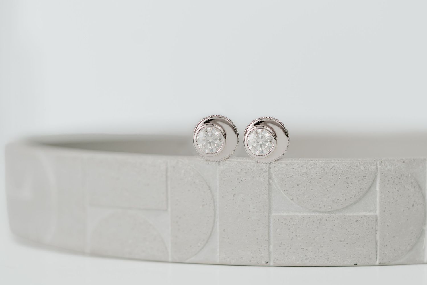 A close-up view of delicate diamond stud earrings in a bezel setting on a light gray background.