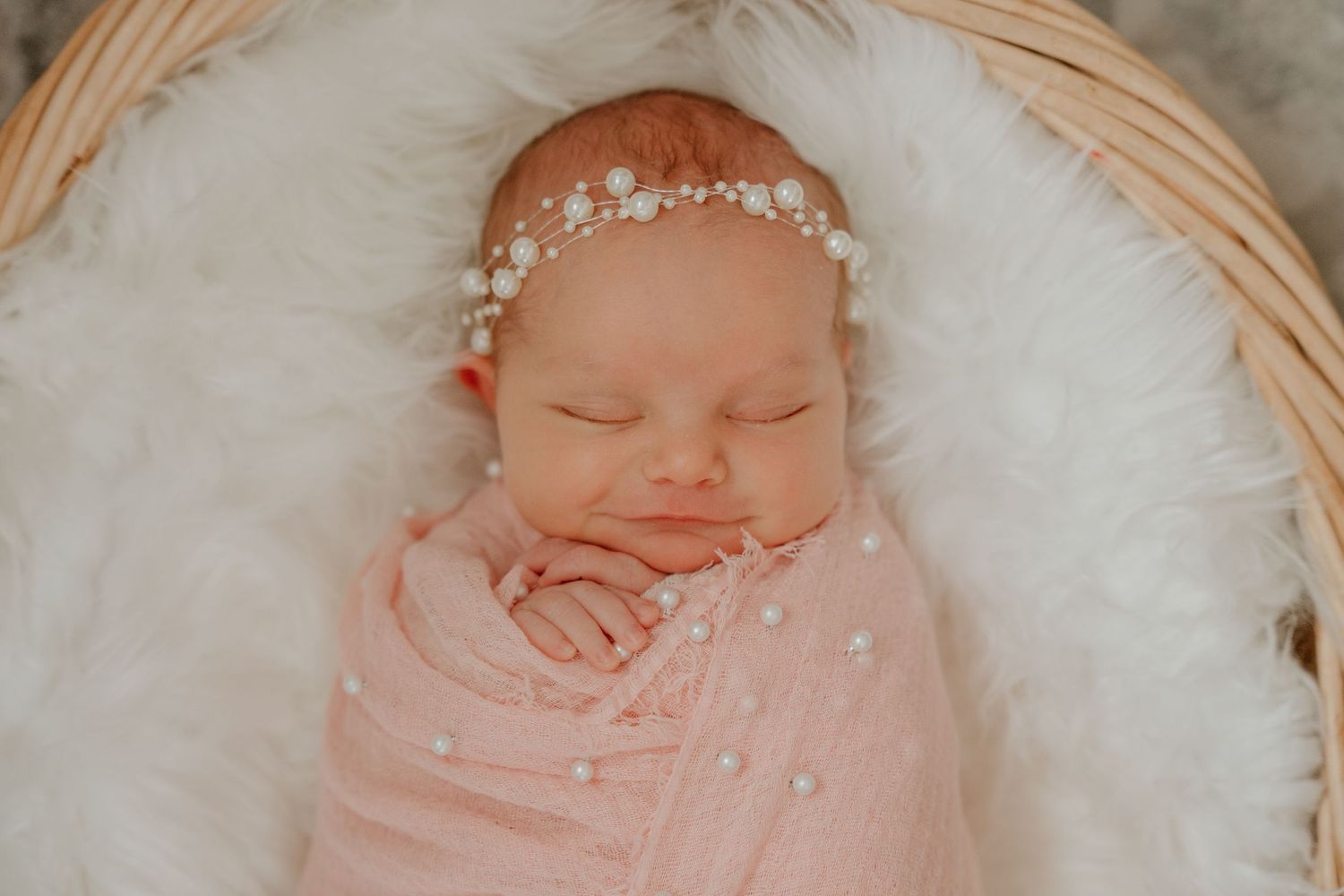 A sleeping newborn wrapped in pink swaddle with delicate pearl headband rests on white fur in a basket.