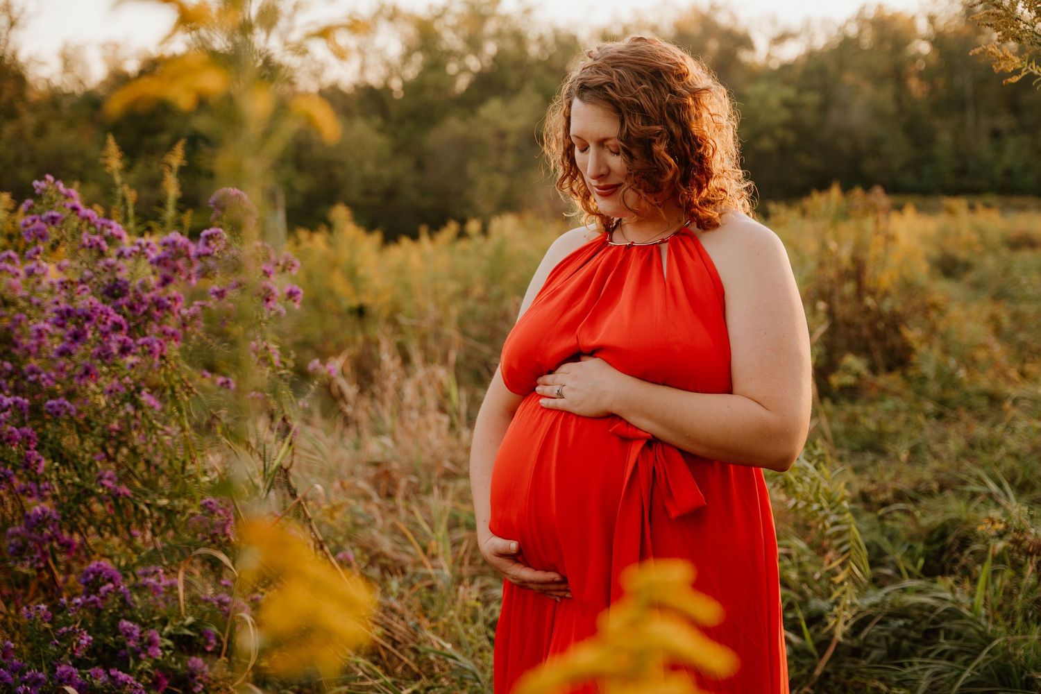 A pregnant figure in a flowing red dress stands among wildflowers at sunset in a natural outdoor setting.