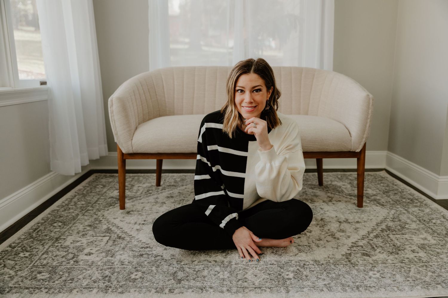 A person in a black sweater sits on a patterned area rug in a bright, modern living room setting.