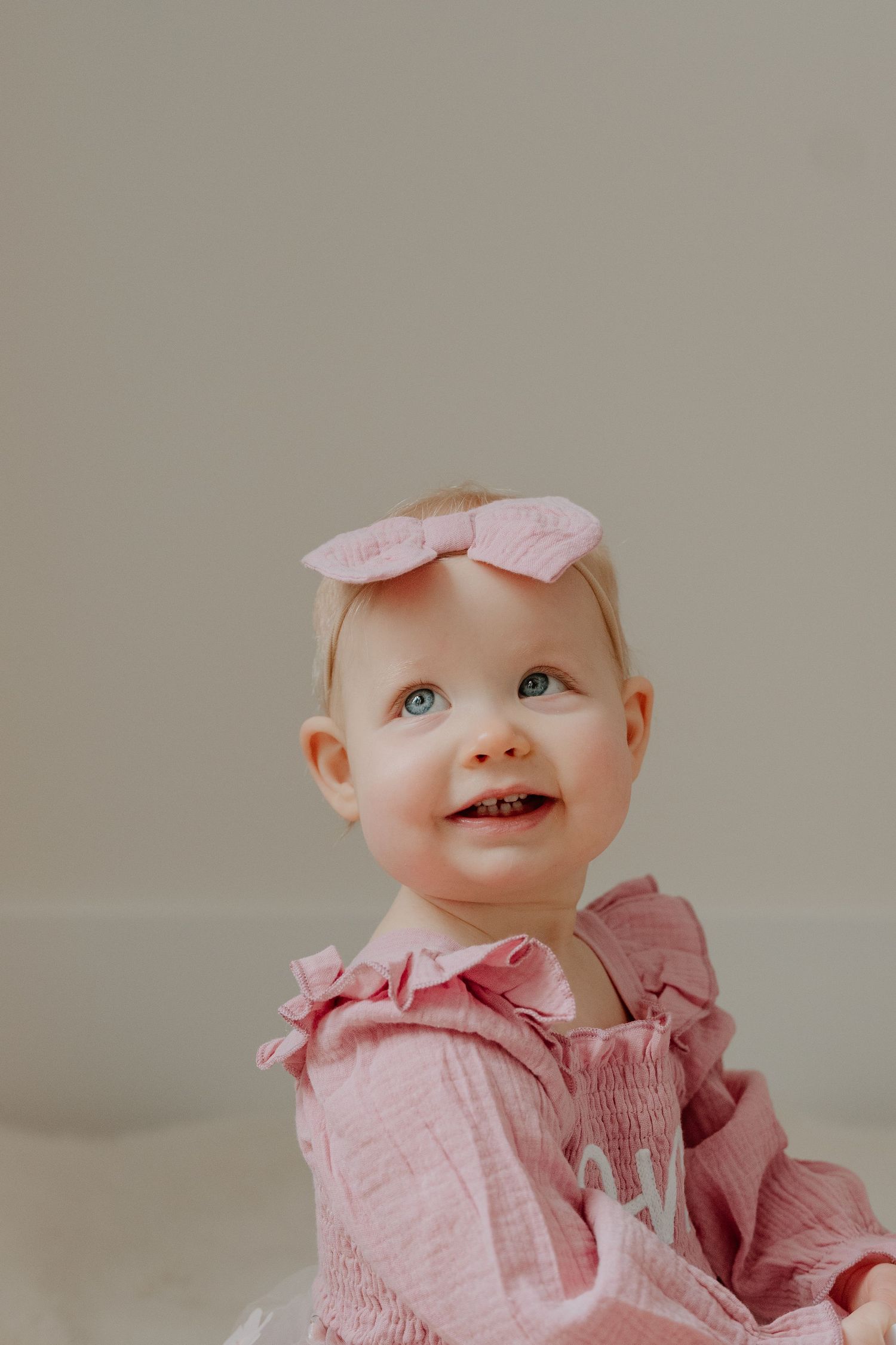 A baby in a pink ruffled dress smiles sweetly while looking up against a neutral background.