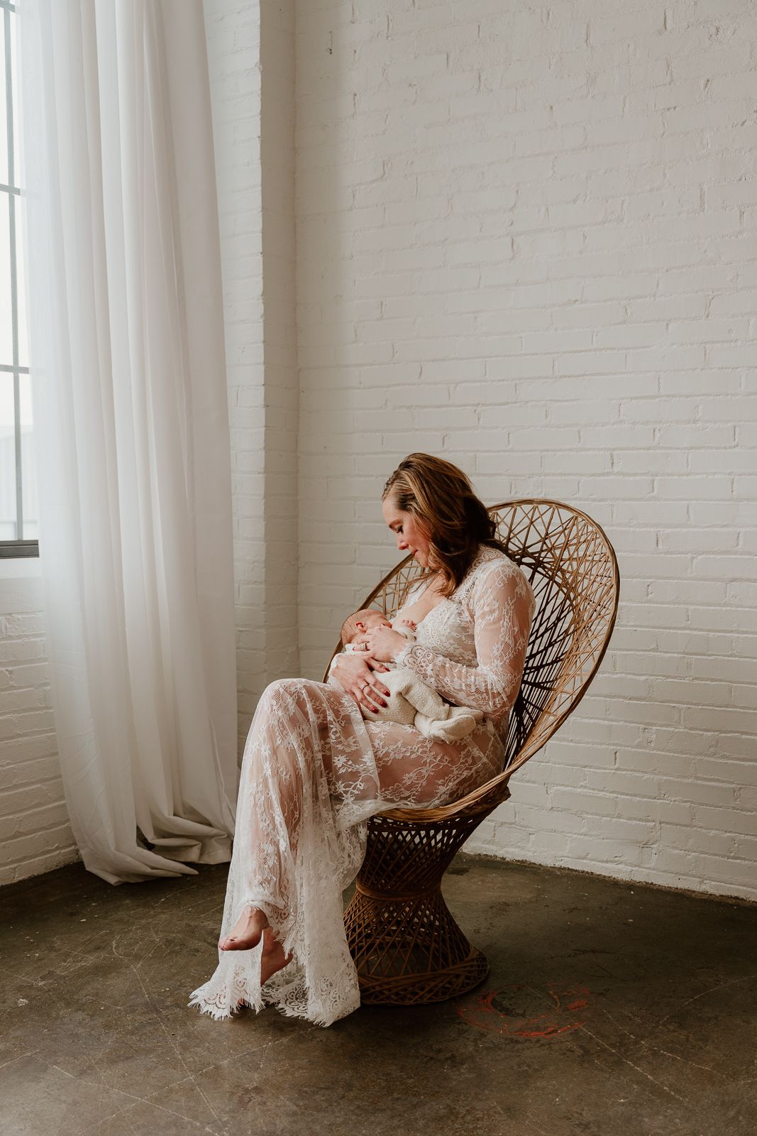 Expecting mother in white lace gown sits peacefully in wicker peacock chair by window in minimalist white room.