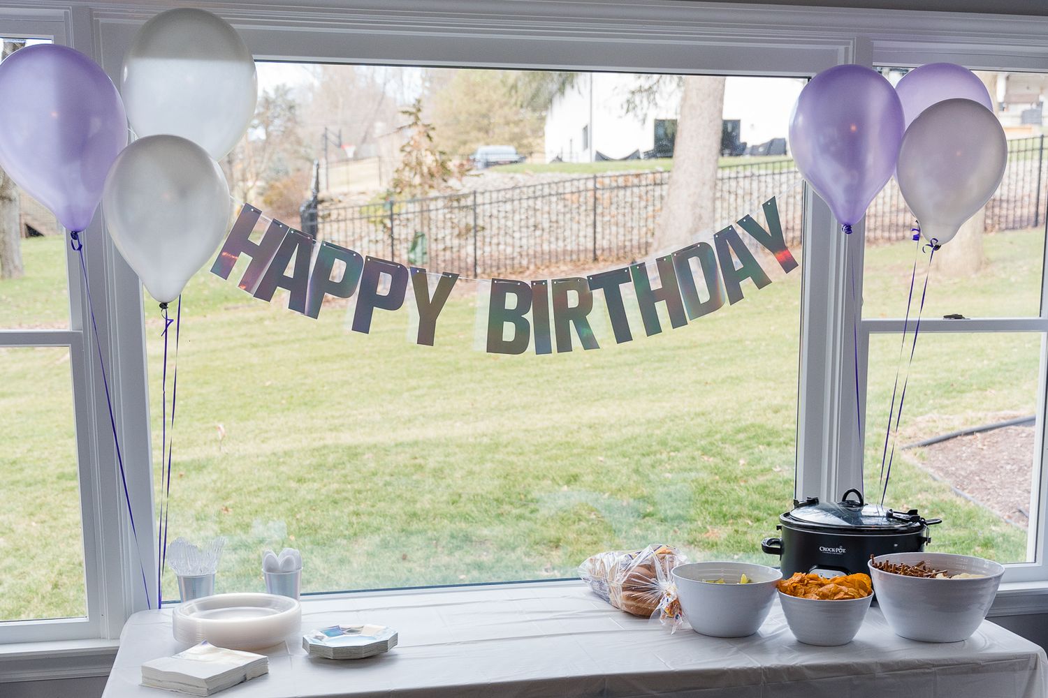 Happy Birthday banner with purple balloons decorating a window with party snacks on display.