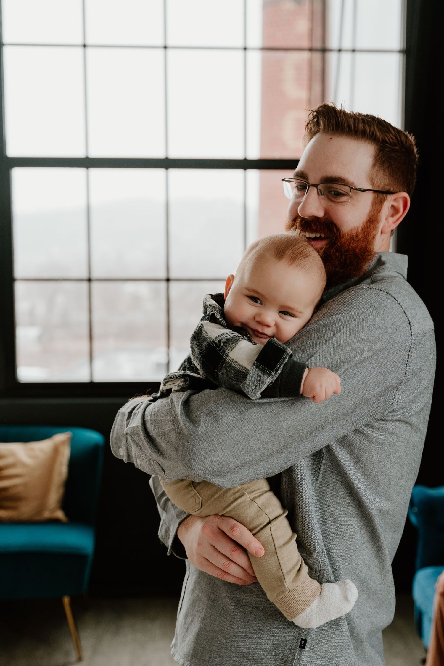 A bearded adult wearing a gray sweater holds a smiling baby in a cozy window-lit room with blue furniture.