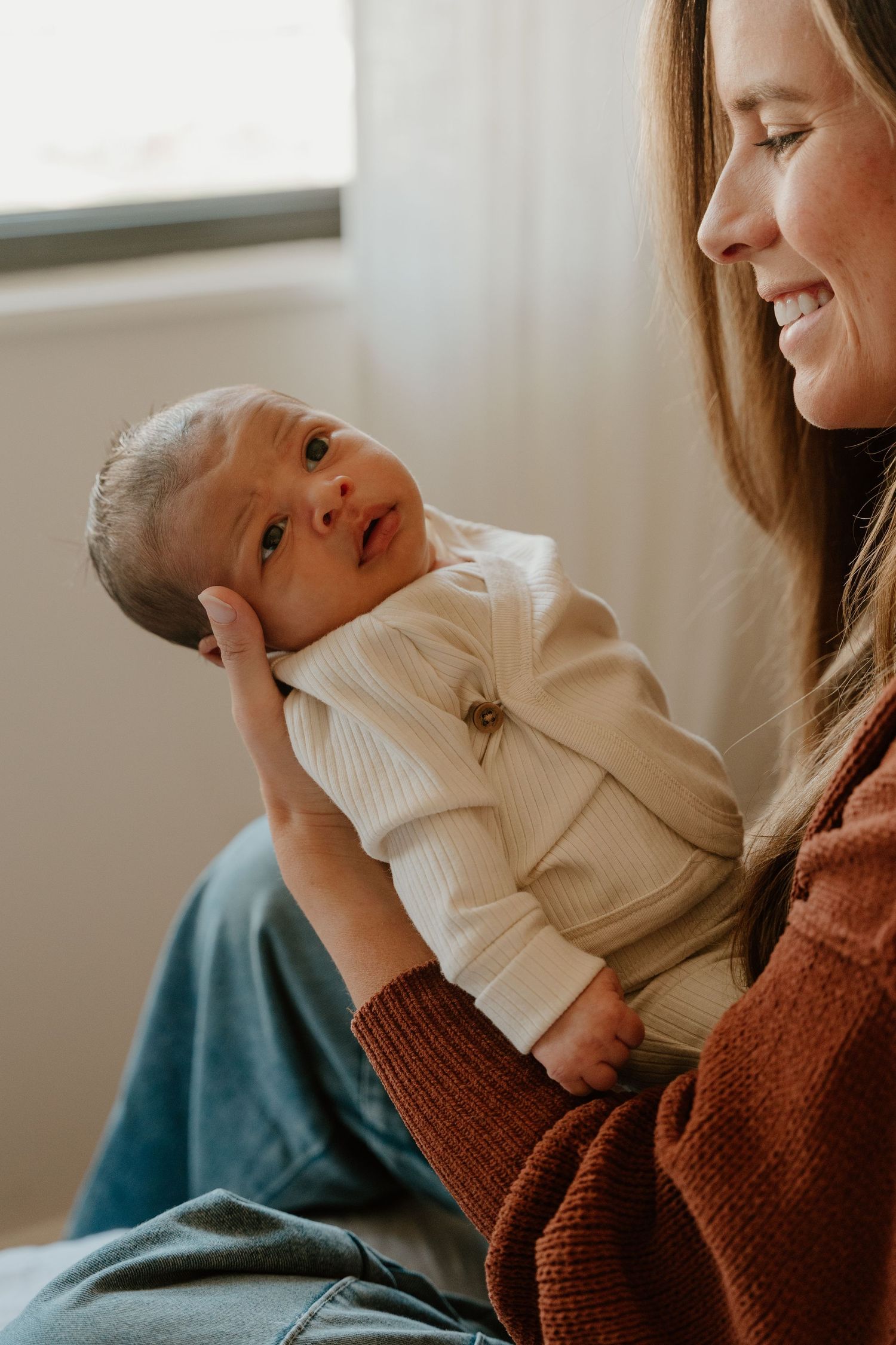 A parent in a brown sweater holds and interacts lovingly with a baby wearing a white turtleneck sweater.