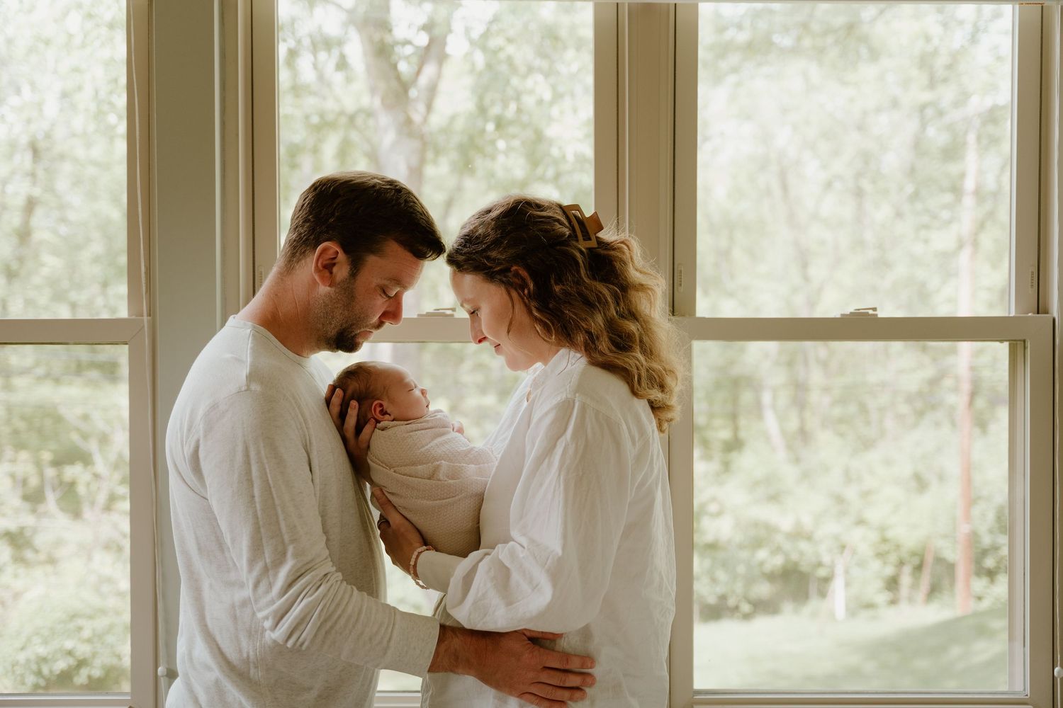 A couple in white outfits share an intimate moment by large windows in a bright, naturally lit room.