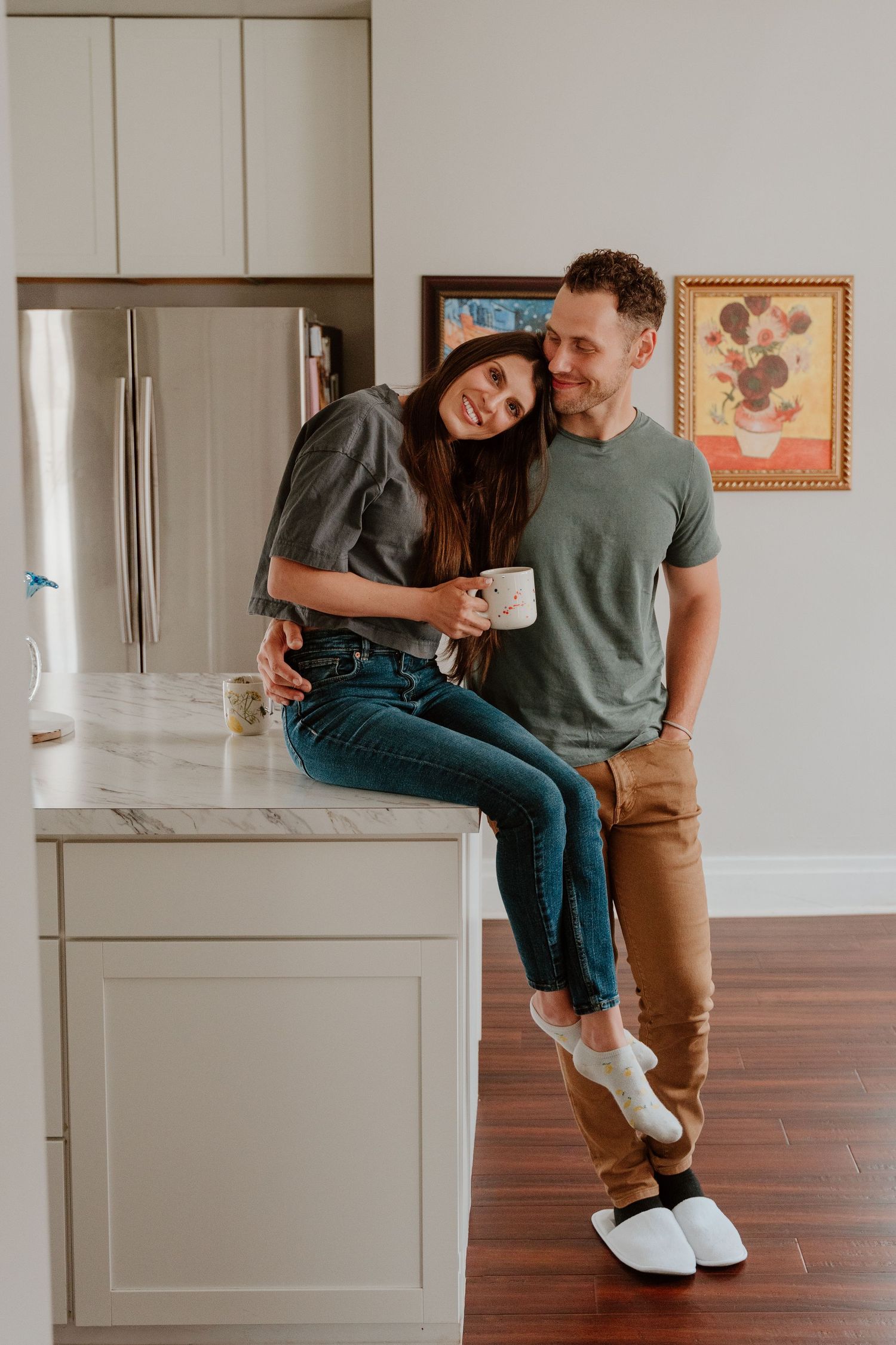 A couple embraces in a modern kitchen while sitting on countertops wearing casual clothing.