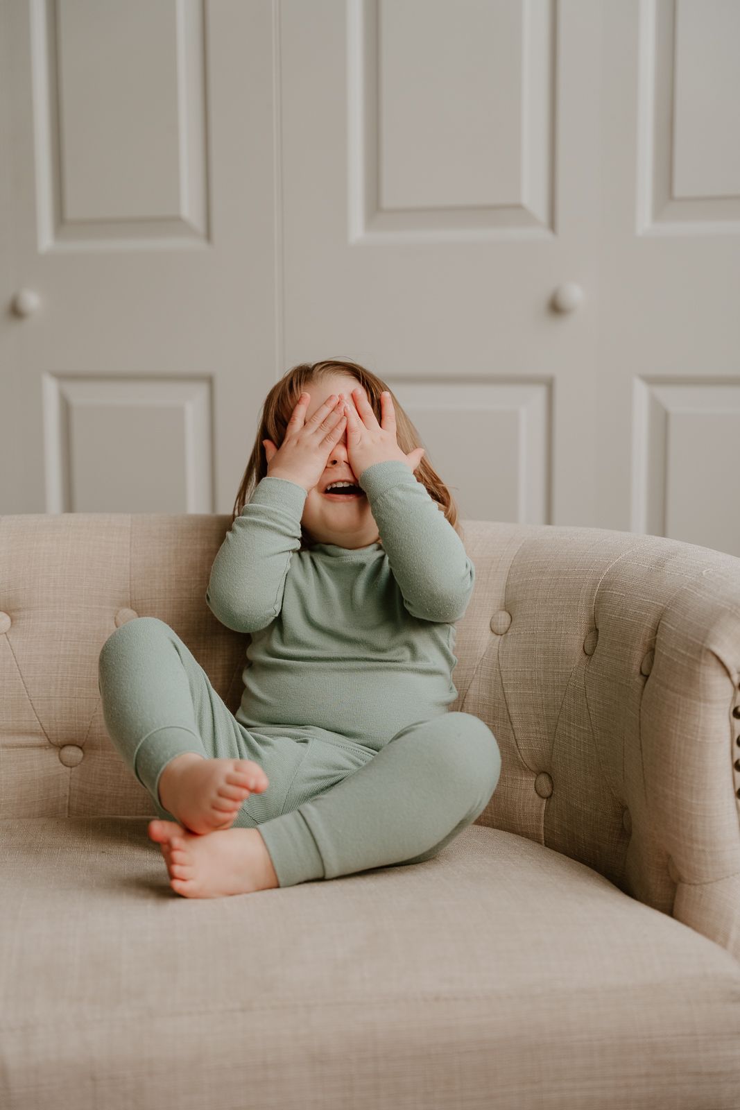 Small child wearing cozy mint green pajamas sits on beige tufted couch while covering face with both hands.