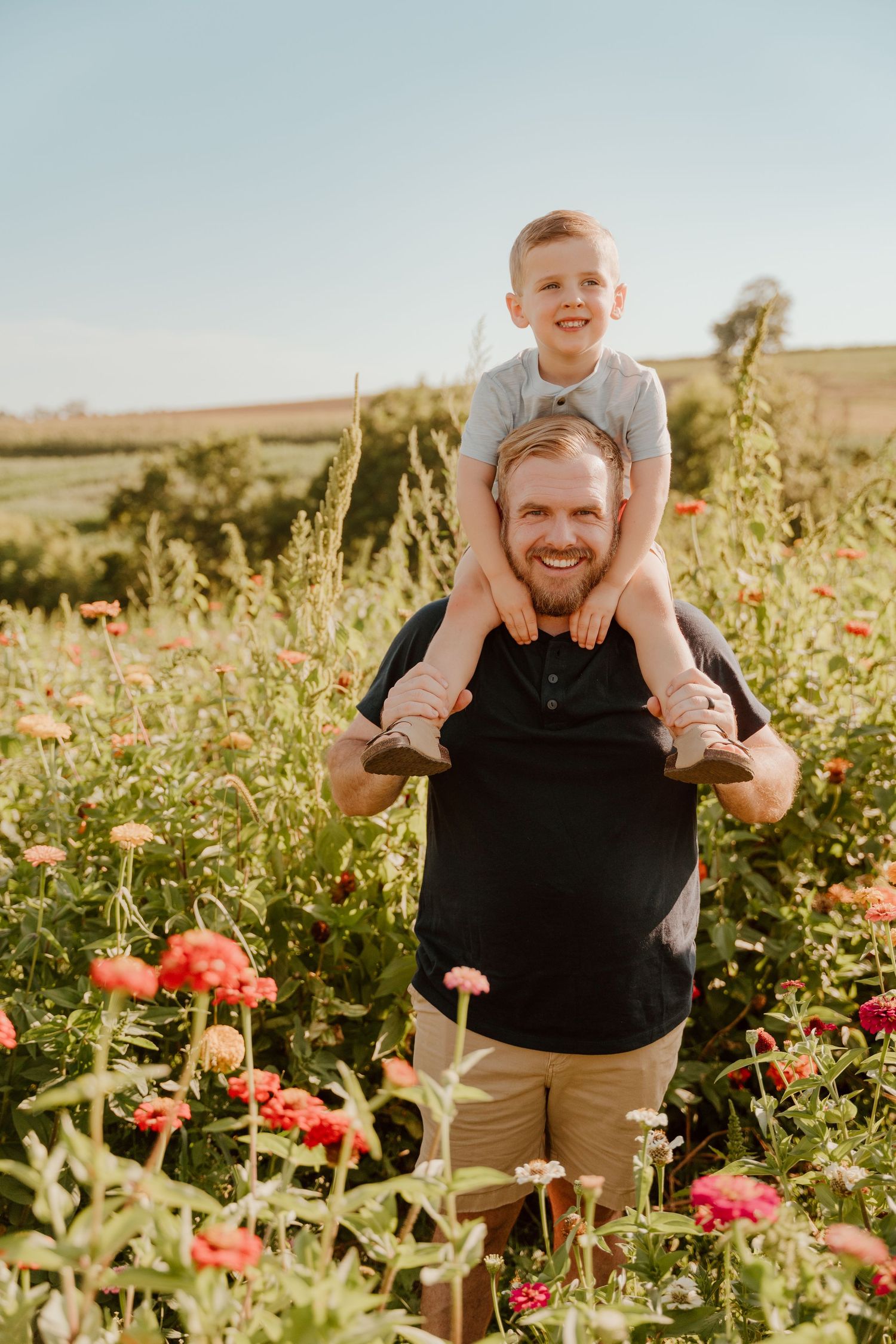 A bearded man smiles while carrying a young boy on his shoulders in a vibrant field of red flowers.