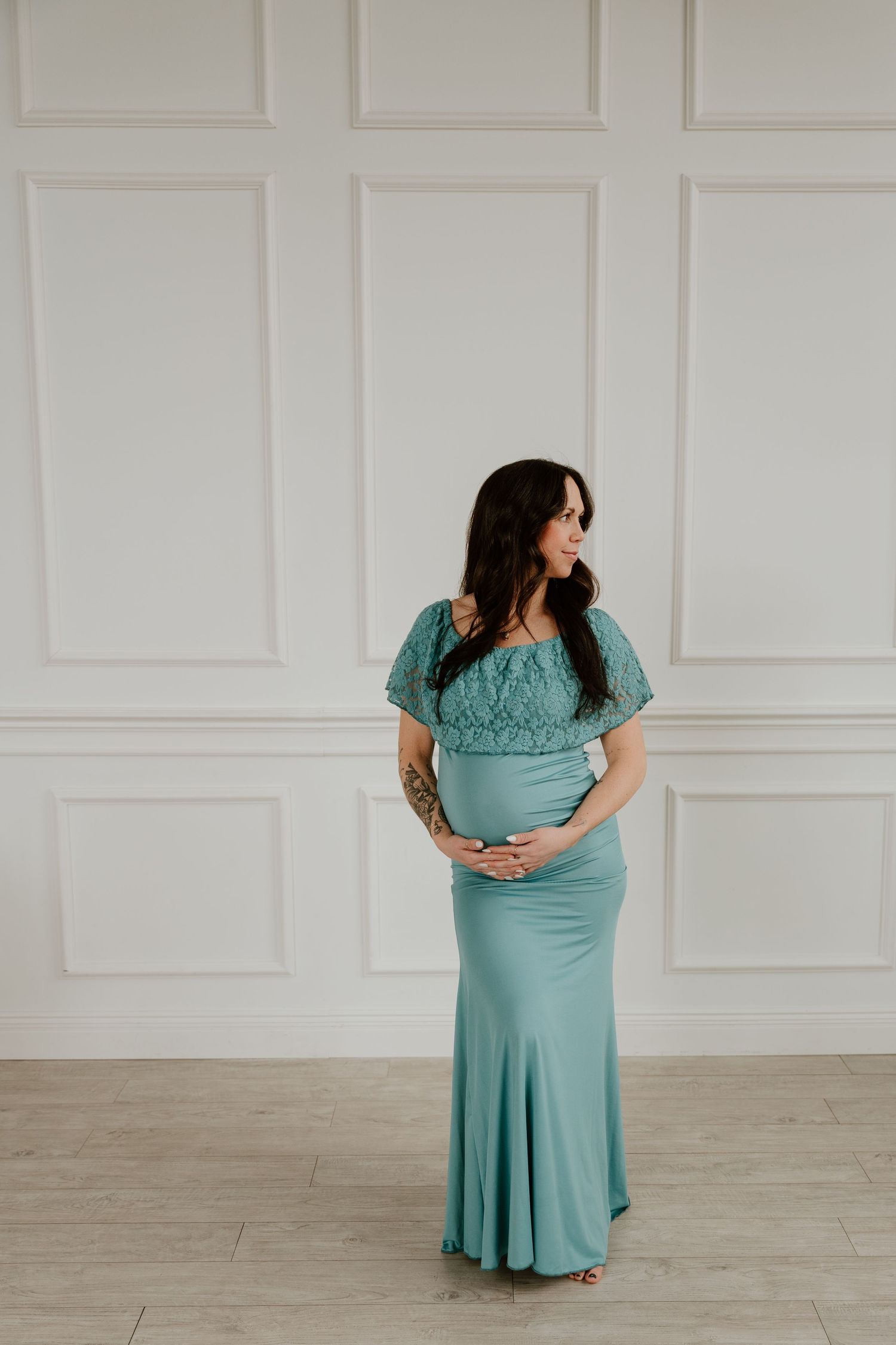 Expectant woman in a flowing turquoise maternity gown poses elegantly against a white paneled wall.