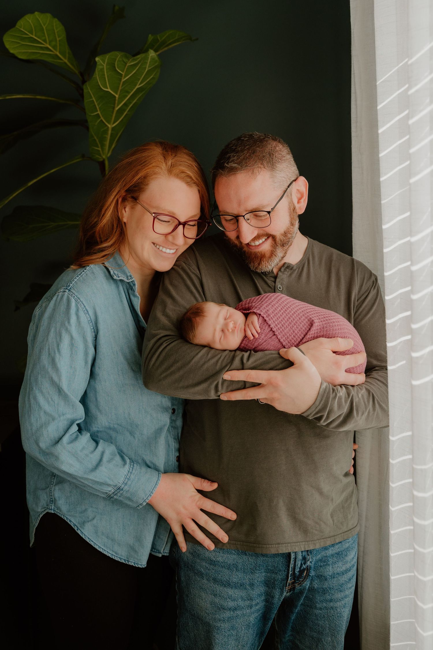A couple in casual attire shares tender moments while holding their newborn baby in a series of intimate indoor portraits.