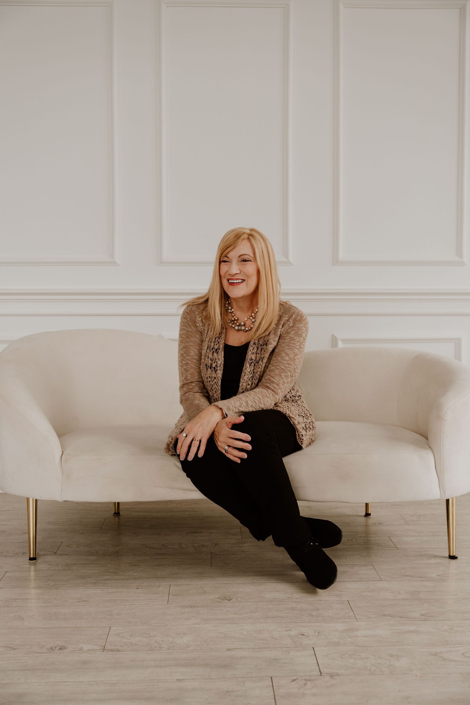 A person in a cozy beige cardigan and black outfit lounges playfully on a white curved sofa against a light gray wall.