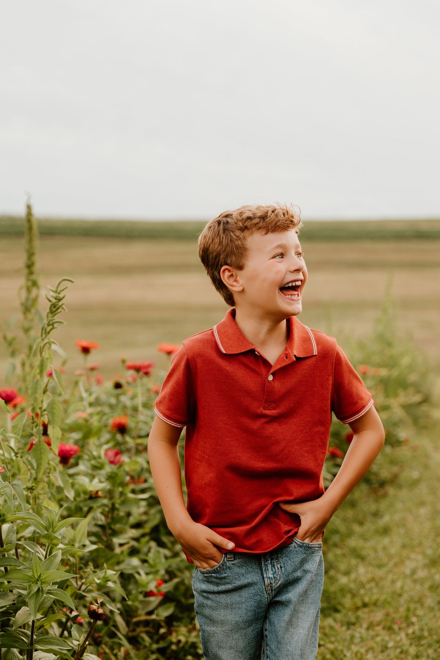 A laughing boy in a red polo shirt and jeans stands among colorful flowers in an open field.