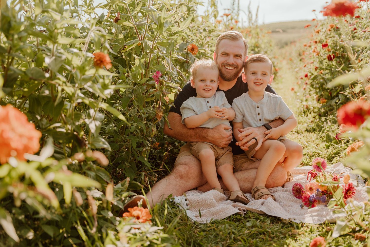Father sits with two young sons on a blanket in a vibrant flower field, sharing a warm family moment outdoors.