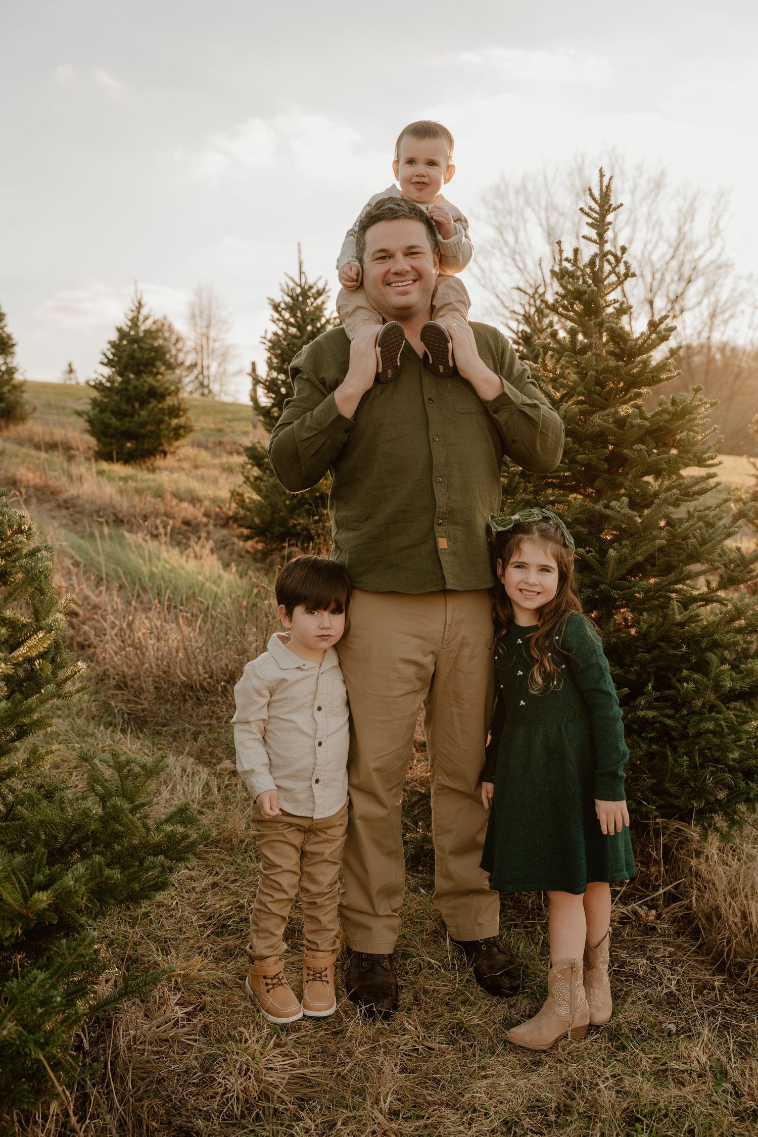 Family portrait in casual attire standing among evergreen trees on a grassy hillside at sunset.