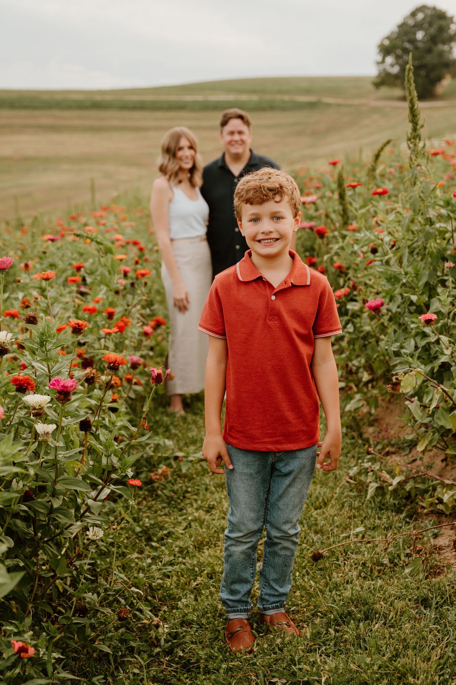 A smiling boy in a red polo shirt stands in a colorful flower field with a couple posing behind him.