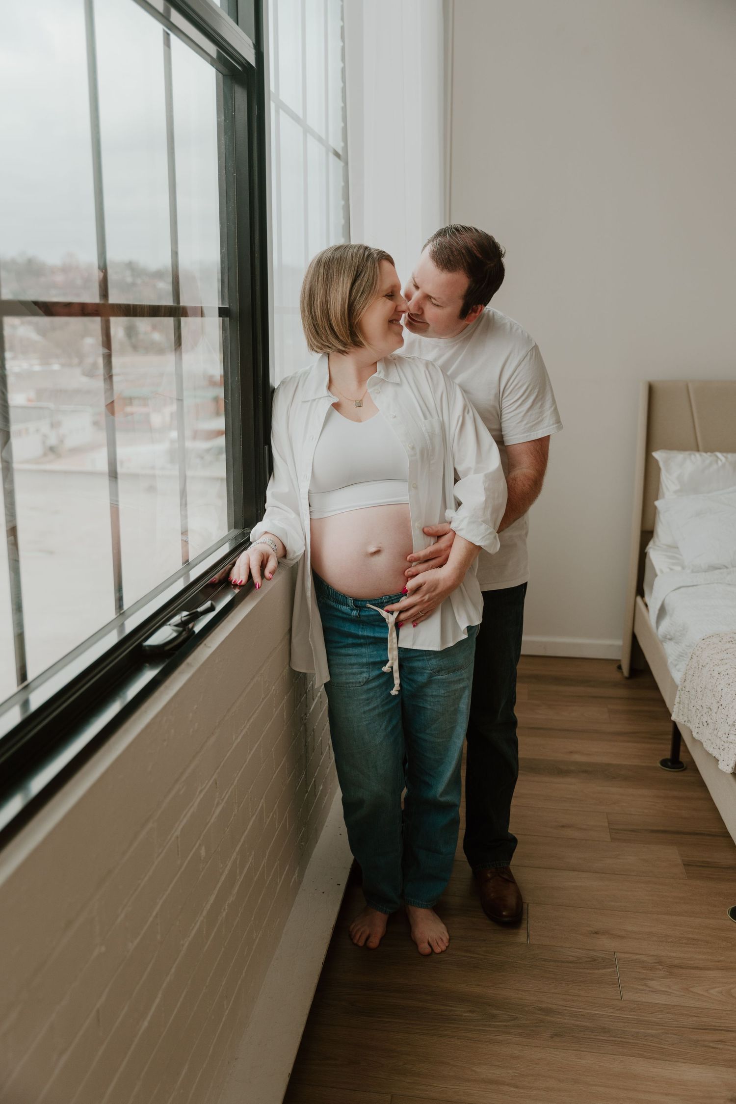 A romantic couple embraces near a window in a minimalist room with hardwood floors and white walls.