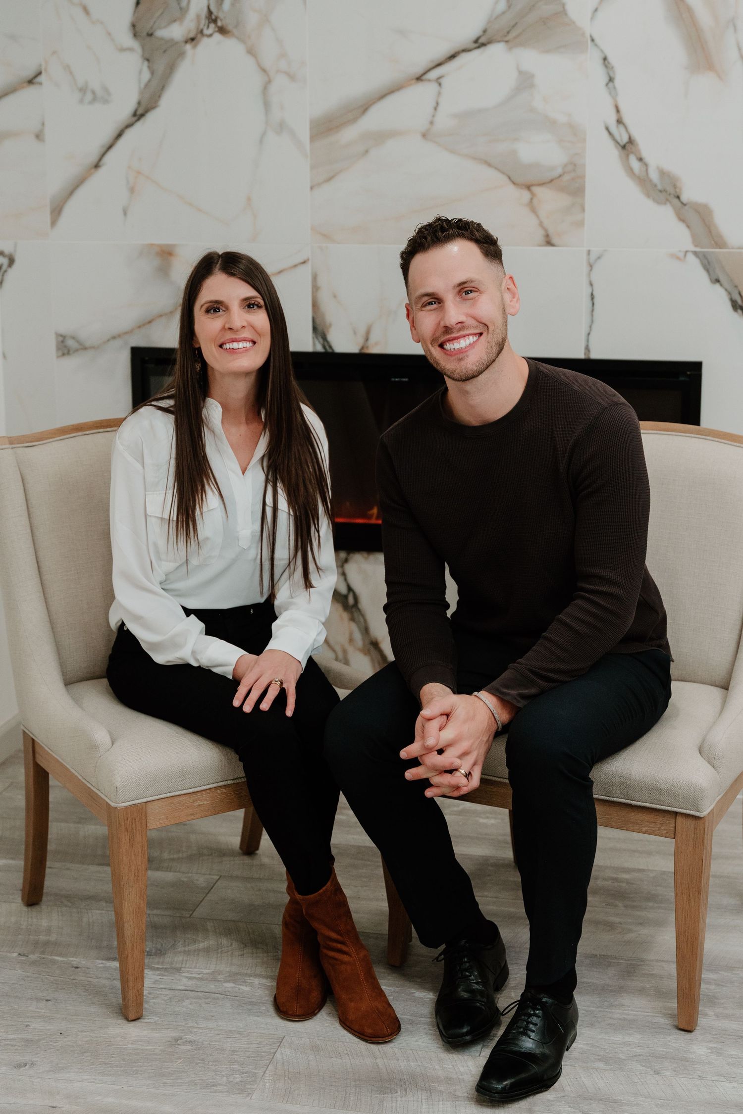 A couple sits together on cream-colored chairs against a marble wall with a fireplace in the background.