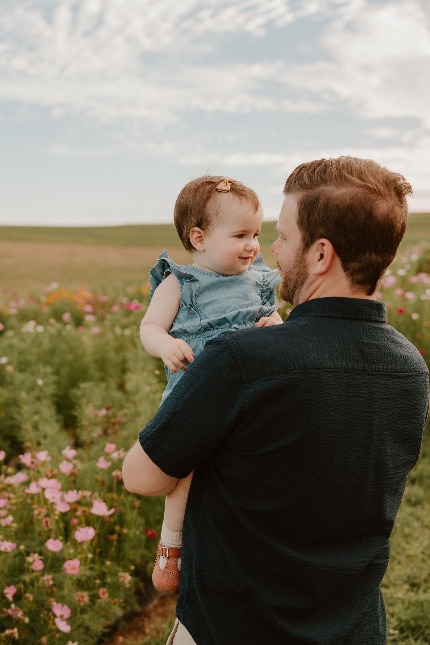 A father holds his smiling baby girl in a blue dress among colorful wildflowers in a scenic field.