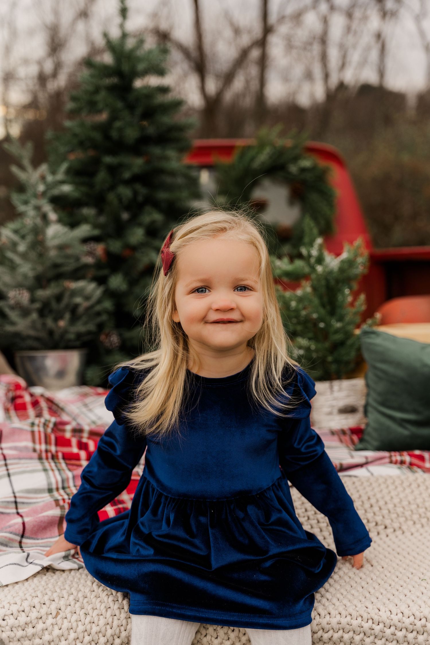 Sweet winter photoshoot with blue velvet dress and evergreen trees in red truck bed backdrop.