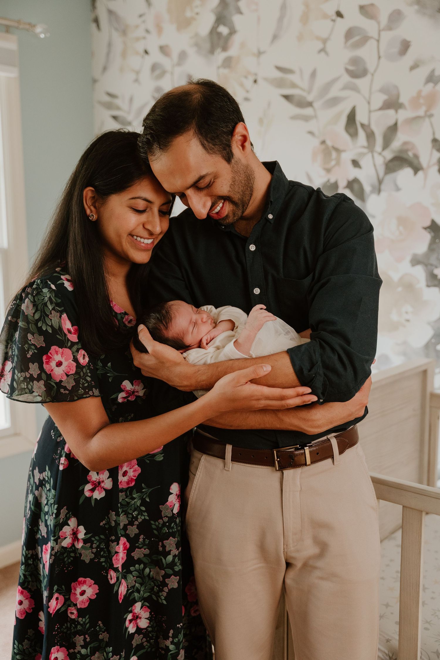 A couple in a floral dress and dark shirt tenderly hold their newborn baby in a sunlit room with floral wallpaper.