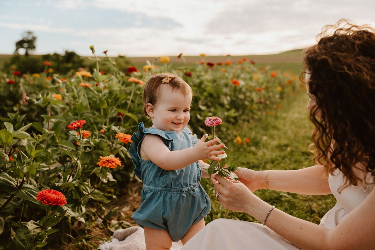 A baby girl in a blue romper holds a pink zinnia flower with her curly-haired mother in a colorful flower field.