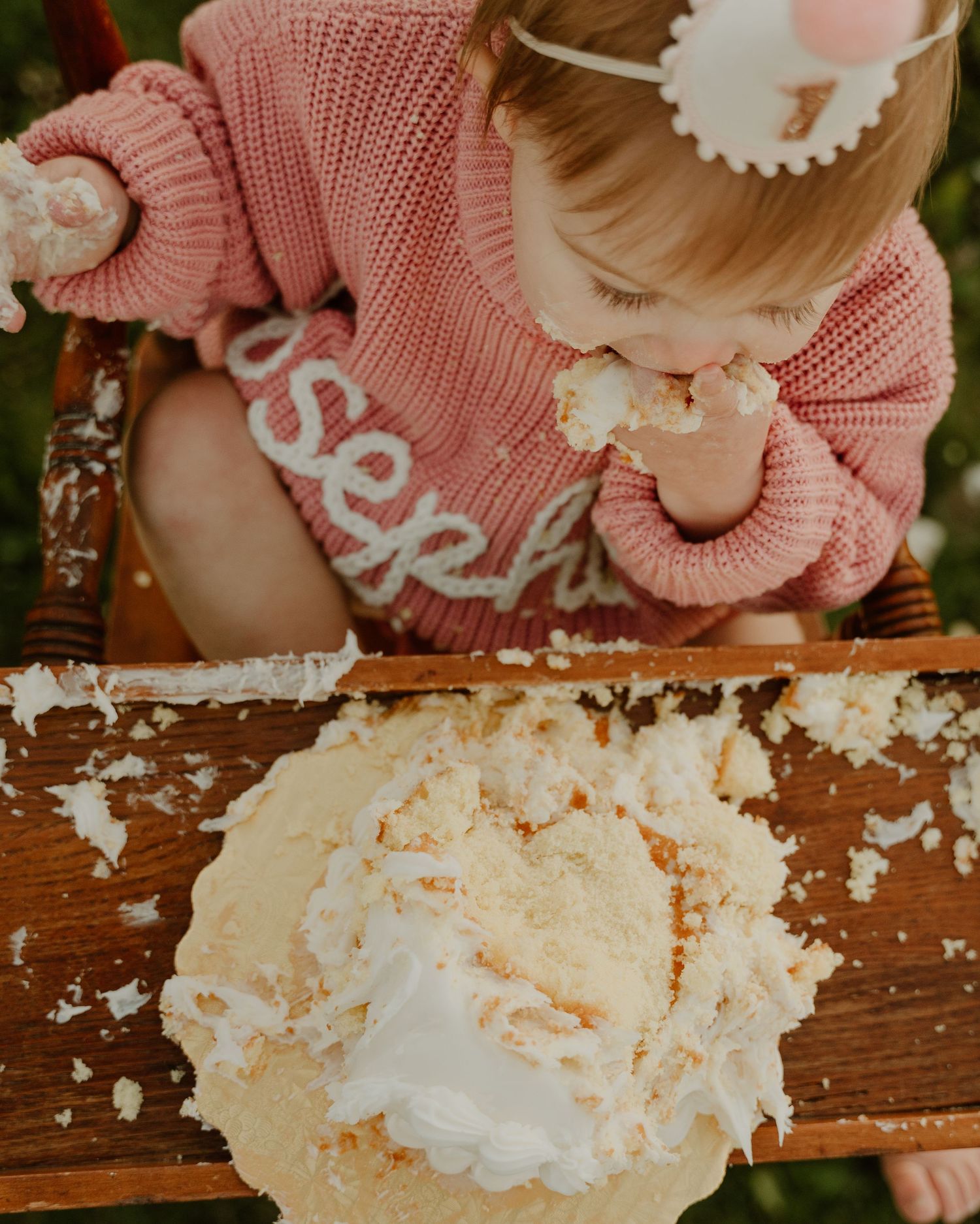 Baby in pink knit sweater smashing and playing with white frosted cake on wooden surface during cake smash photoshoot.