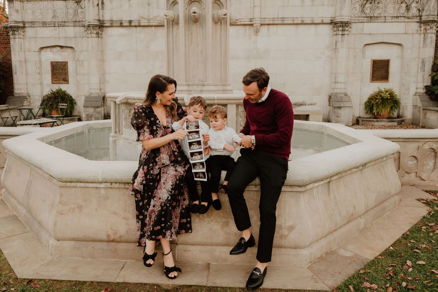 Family sharing playful moment at ornate white stone fountain in garden setting.