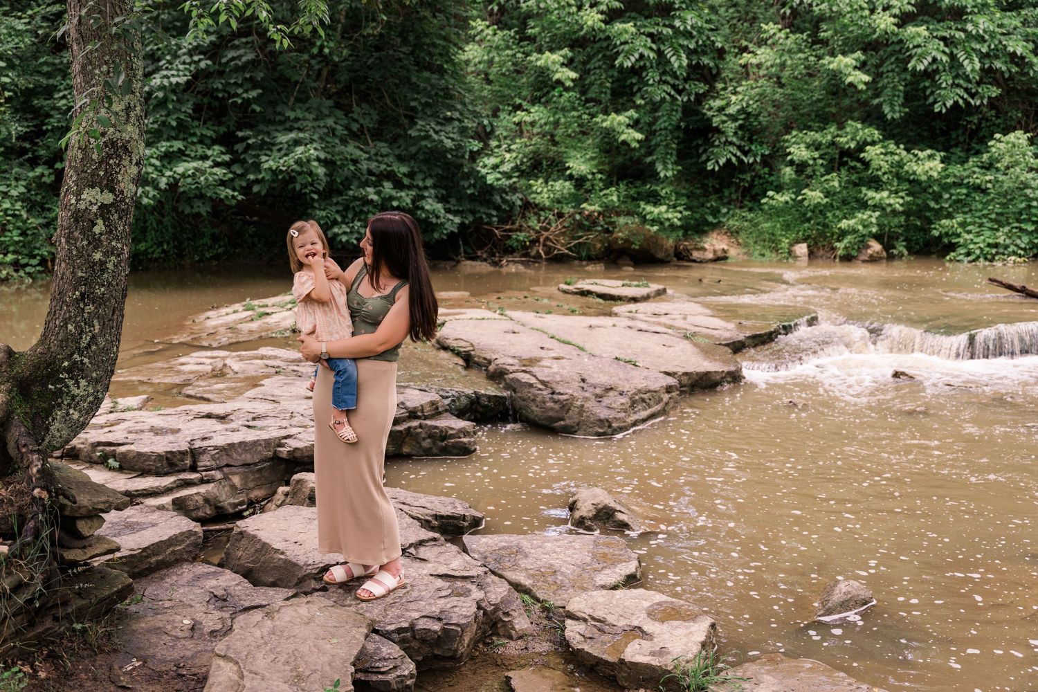 Two people standing on rocky outcrop near flowing river surrounded by green forest.