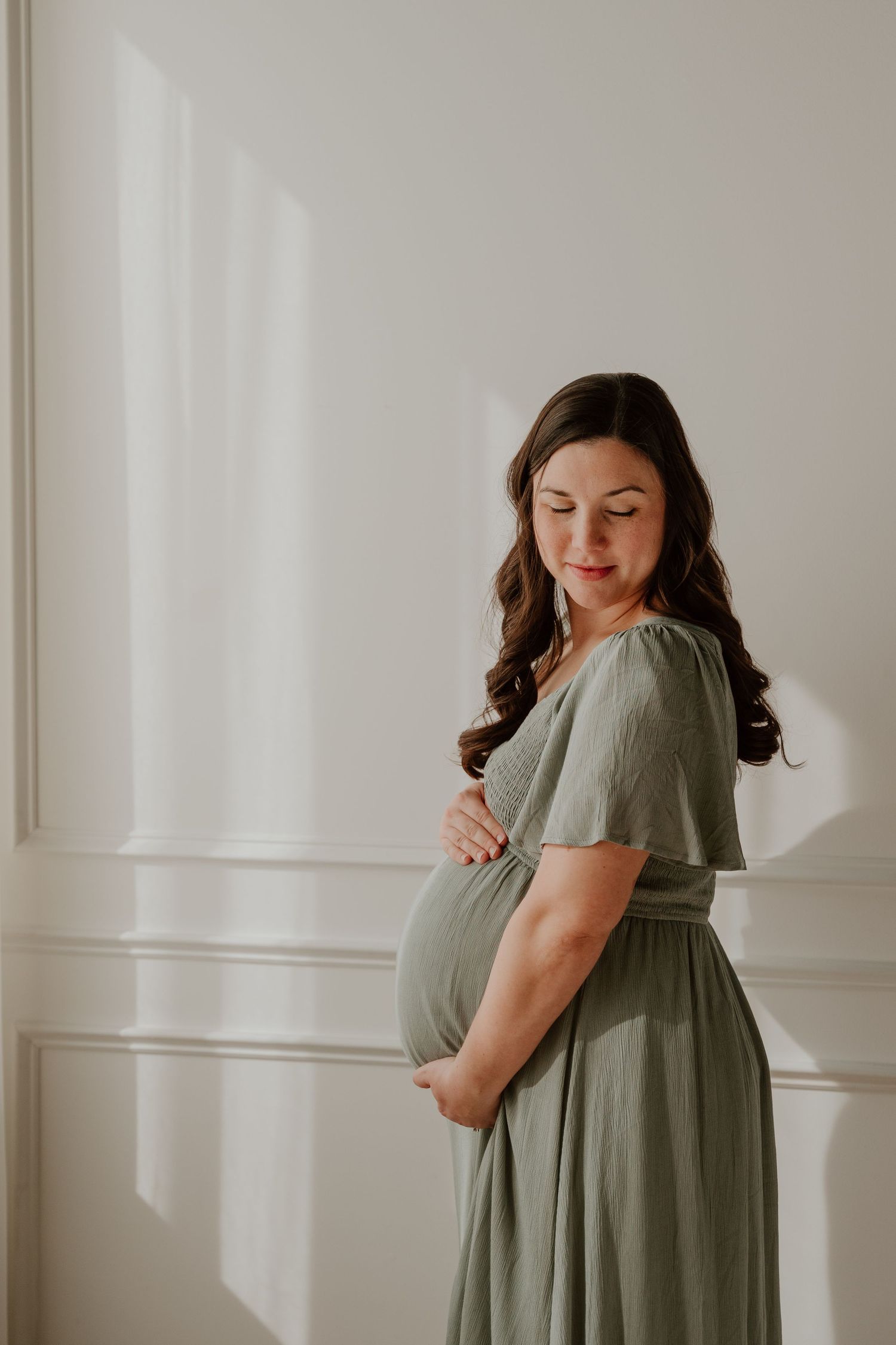 A woman in a sage green flowing maternity dress stands in profile against a white wall with dramatic window shadows.