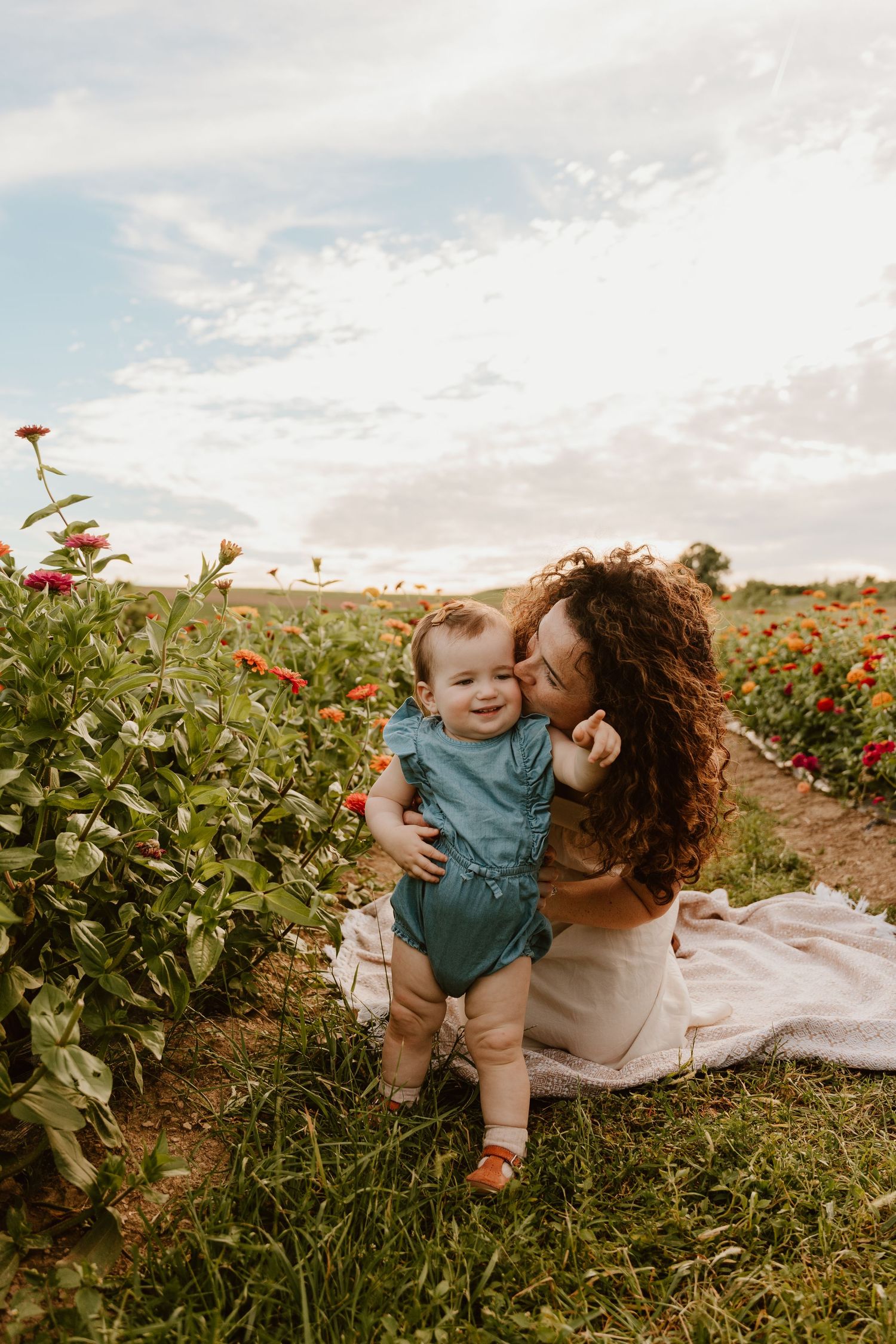 A curly-haired mother kisses her smiling baby girl in a blue romper while sitting in a vibrant flower field.