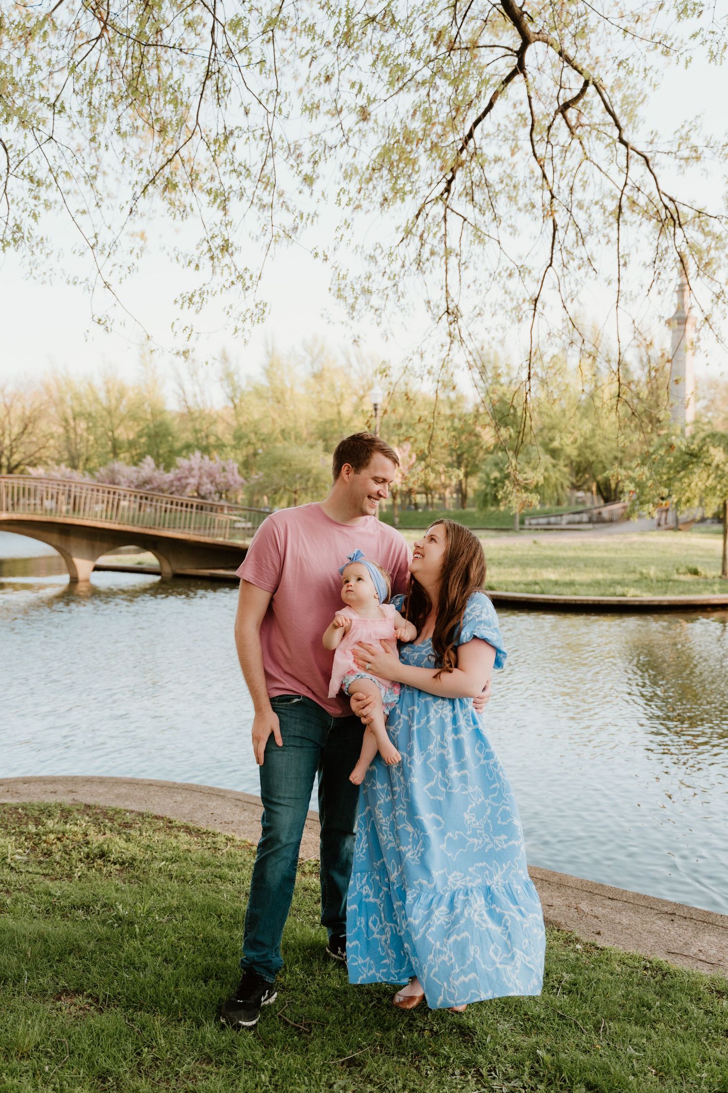 Family standing together by lake with bridge and spring trees in background.