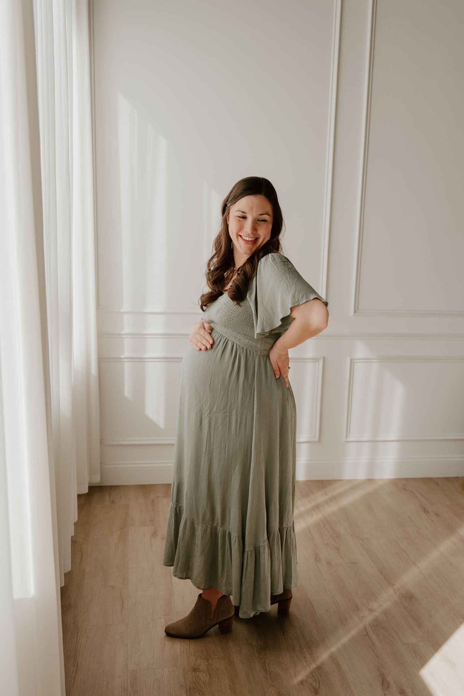 A woman poses in a sage green maxi dress with ruffled hem and brown ankle boots against a white paneled wall.